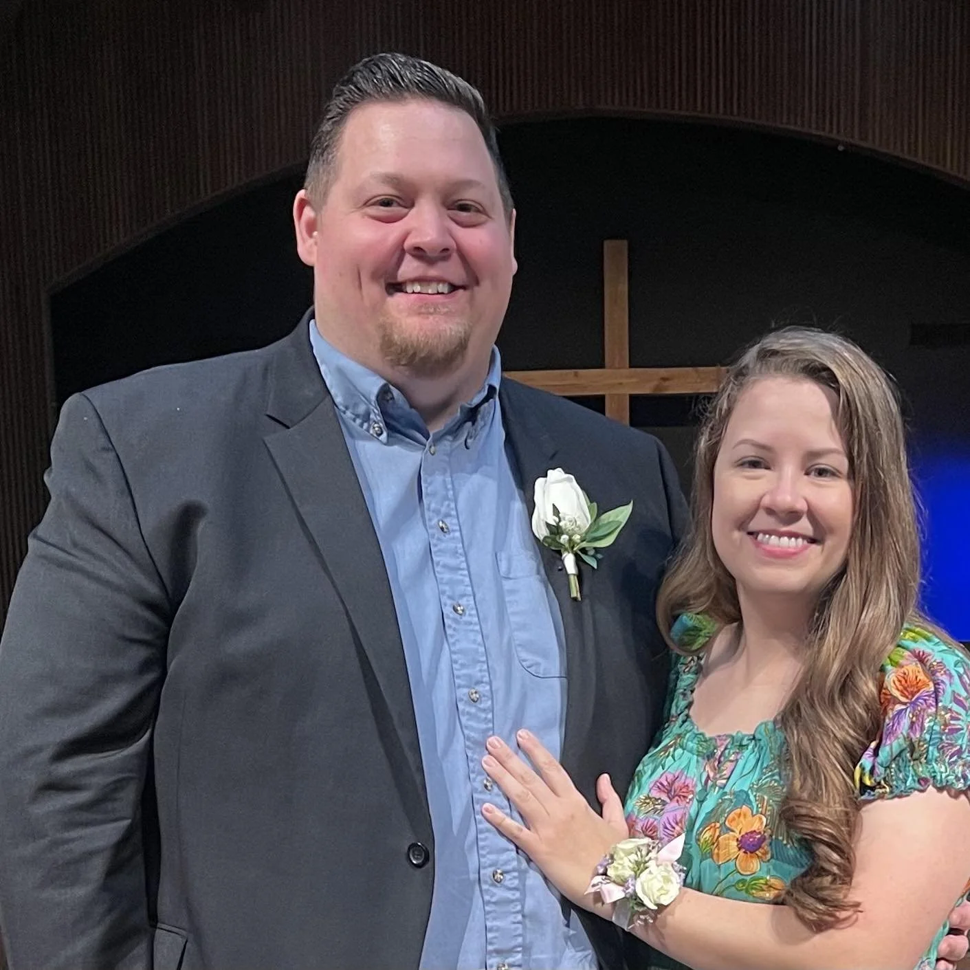 A smiling man and woman standing together inside a church with a cross in the background. The man is wearing a gray suit with a light blue shirt and a white boutonniere. The woman is wearing a colorful floral dress and a matching corsage, with her hand on the man's chest.