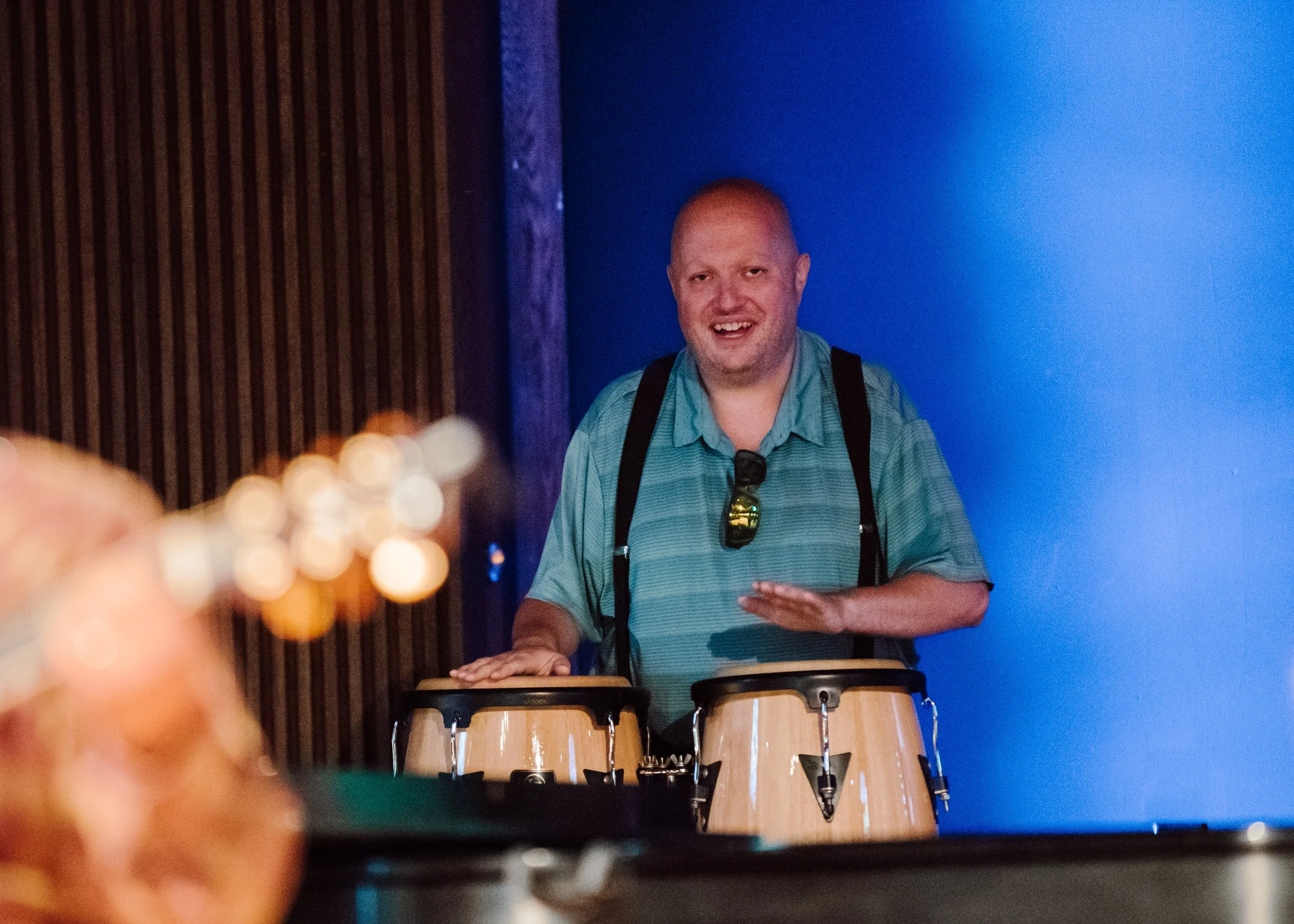 A man with a bald head and a beard, wearing a teal shirt with sunglasses hanging from the collar, is playing conga drums and smiling in front of a blue wall.