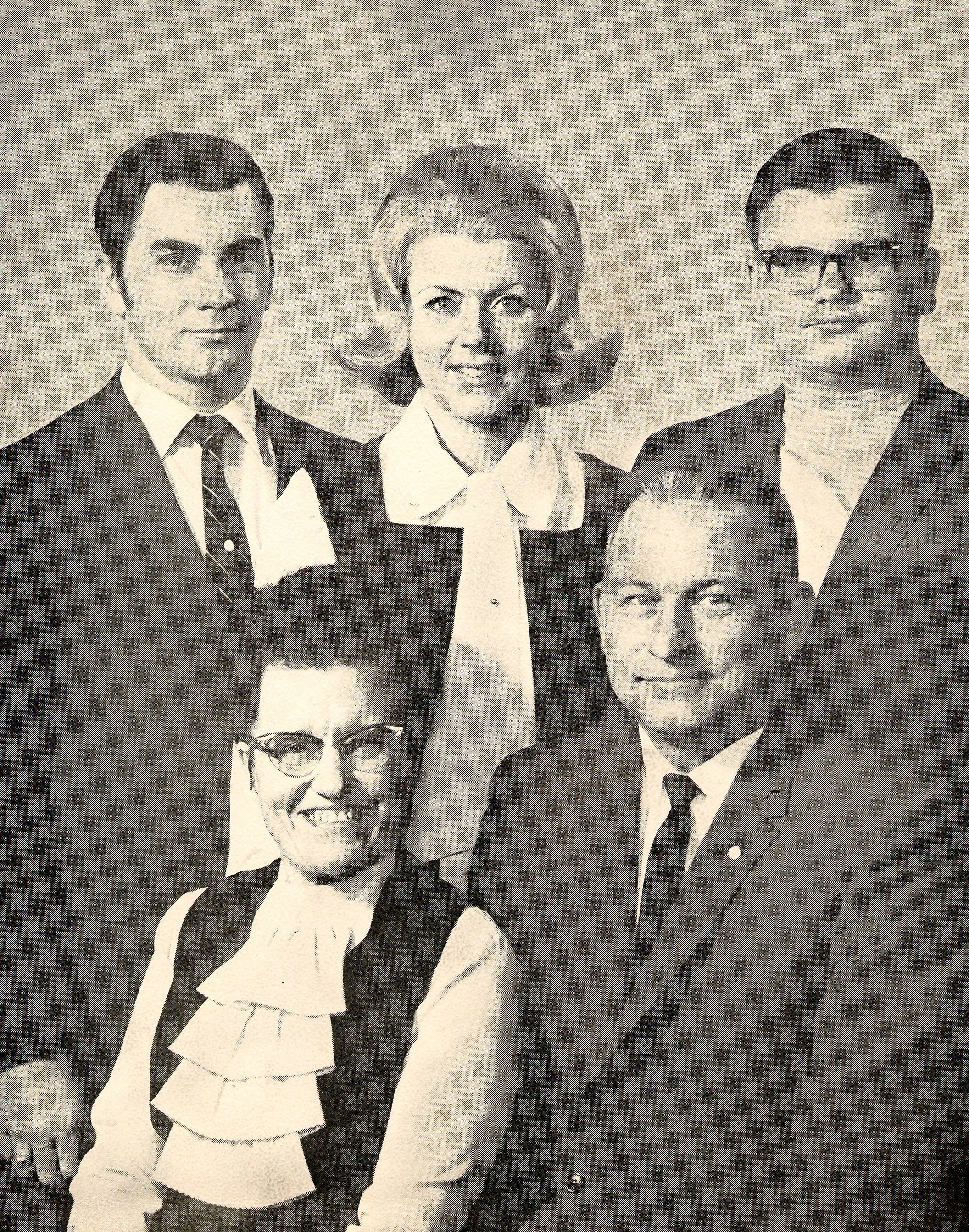 A black and white photo of five adults, three men and two women, dressed in vintage business attire, posing together for a group portrait.