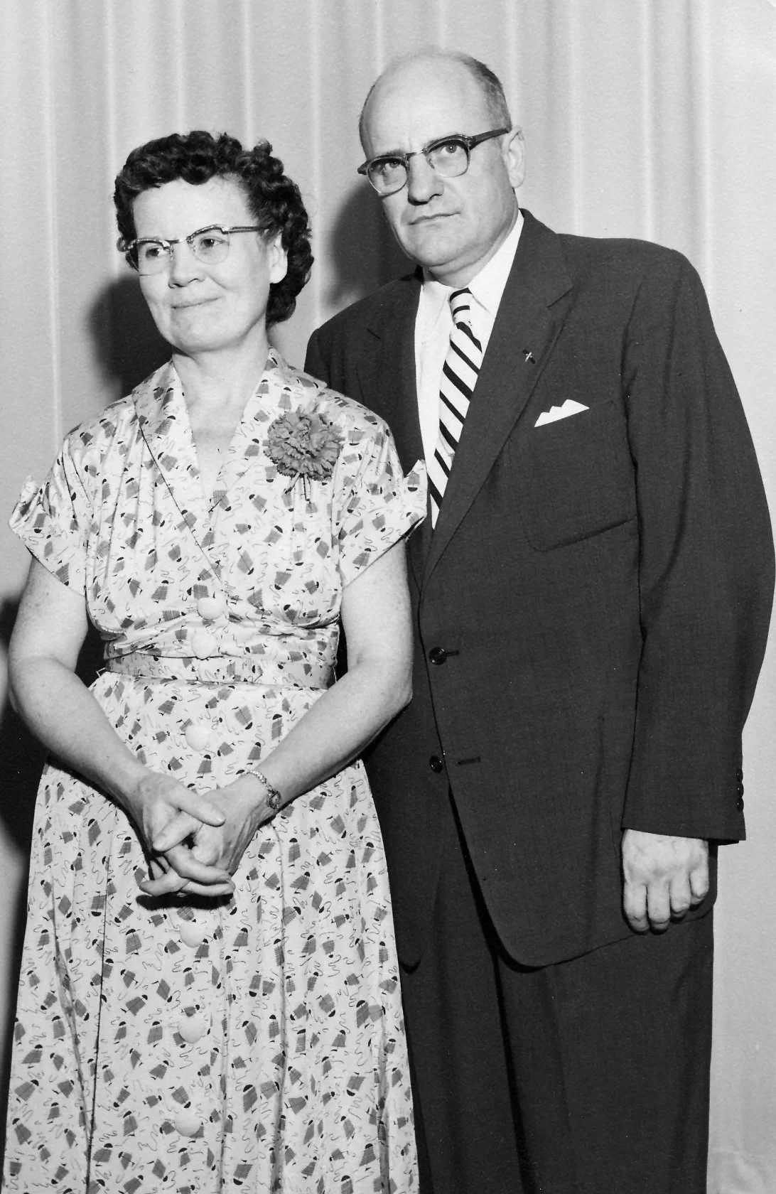 A black-and-white photograph of a woman and a man standing close together at an event. The woman is wearing glasses and a patterned dress with a flower decoration, and the man is wearing glasses, a dark suit, and a striped tie.