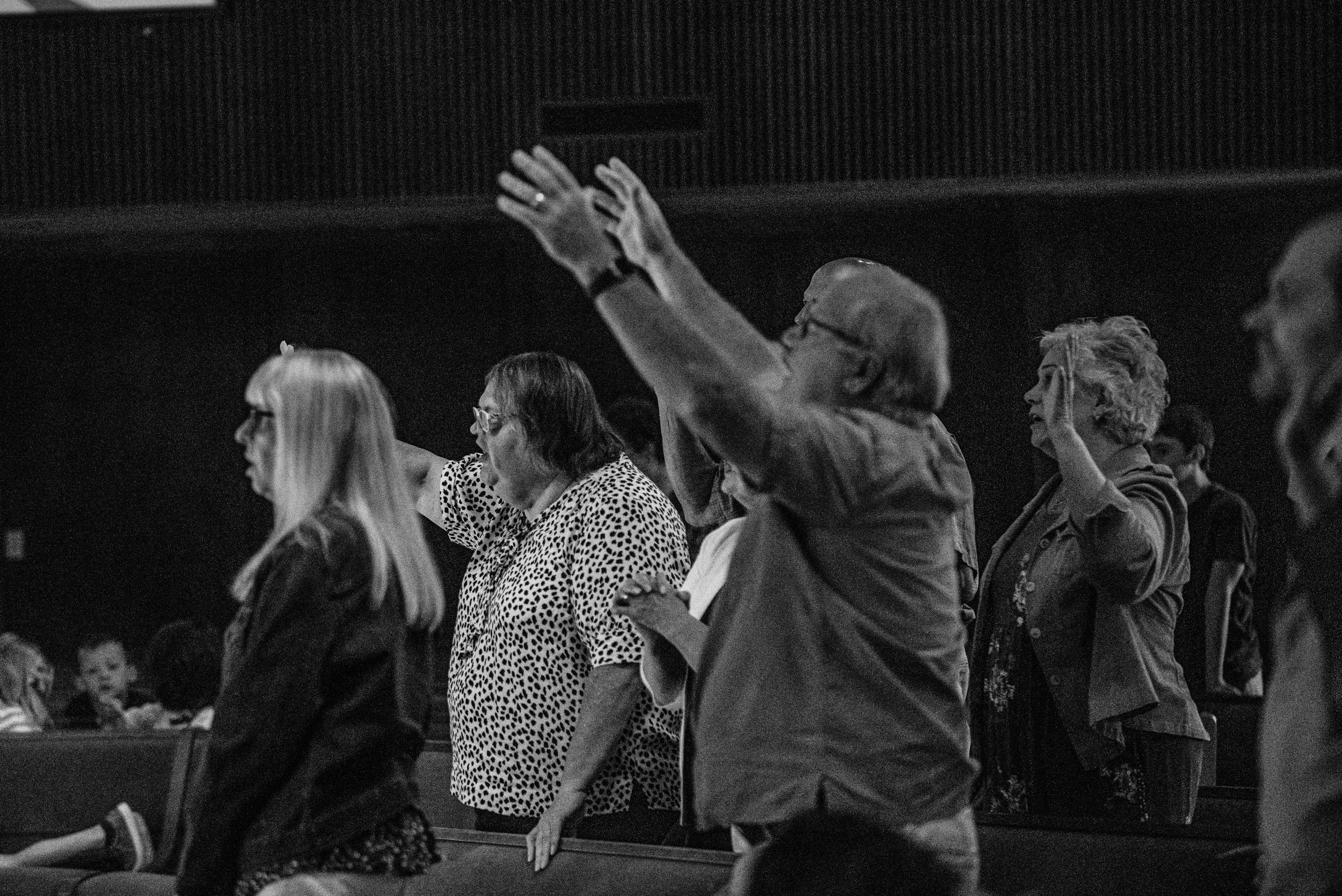 People standing in a church or auditorium with some raising their hands during a worship or prayer service, in black and white.