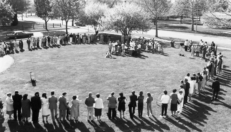People standing in a long line in a park, forming a square around a group of people sitting at tables, with trees and a street in the background.