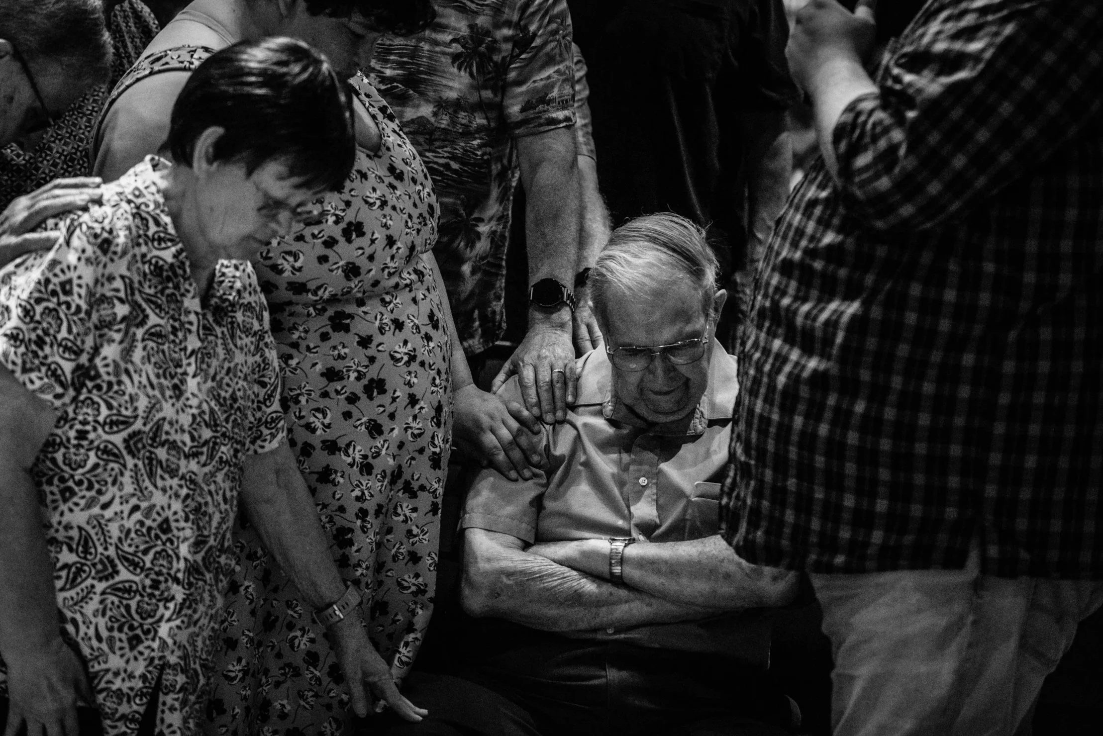 A group of people gathered around an elderly man with glasses, who appears to be in emotional distress or crying, while others support and comfort him.