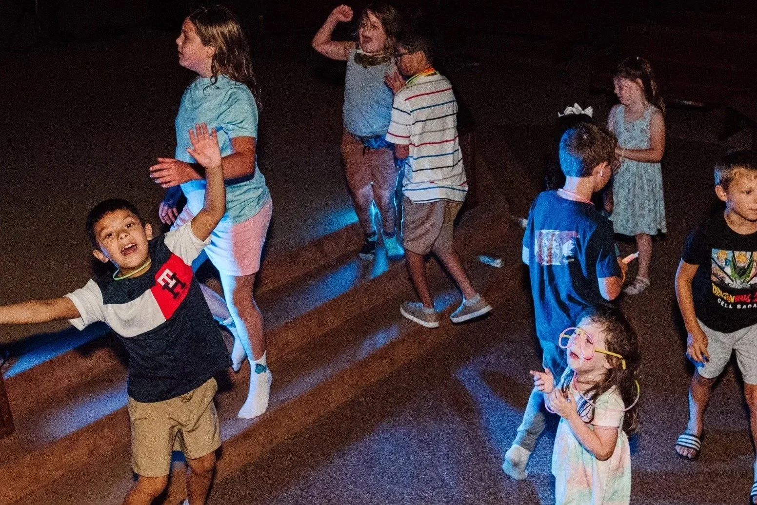 Children dancing and playing indoors, some wearing glasses, with a dark background and colorful lighting.
