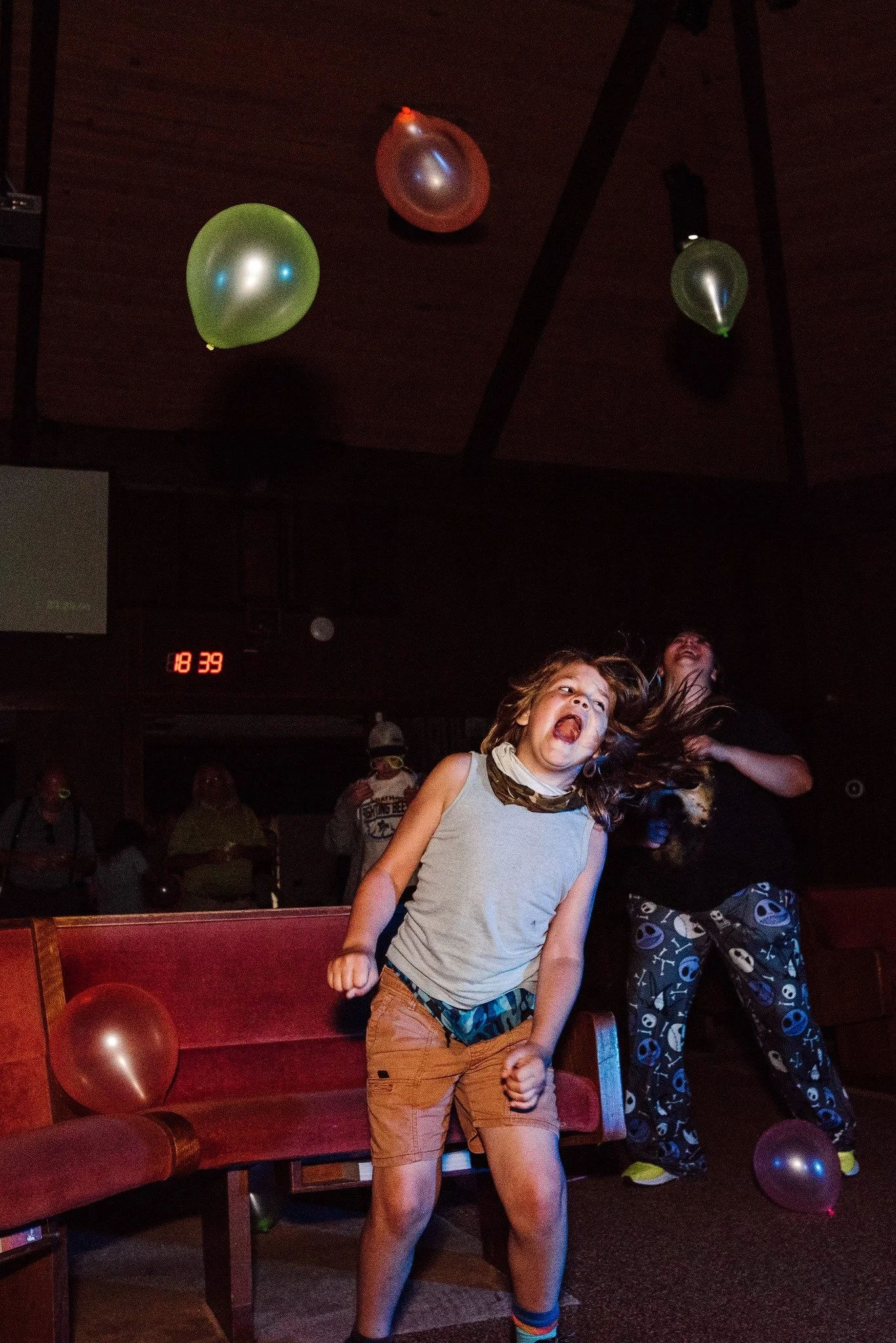 A young girl with brown hair and light skin making an angry face, clenching her fists, in a party setting with balloons hanging from the ceiling and people in the background.