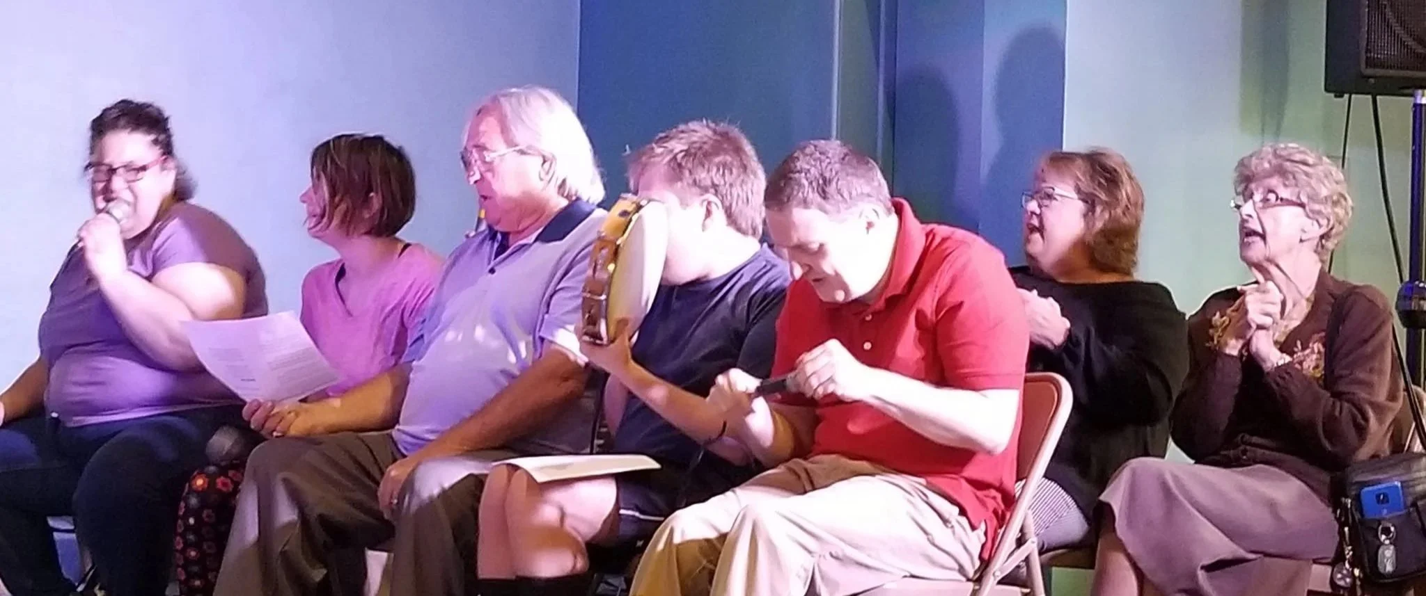 Group of people sitting in a row, some reading papers, others looking at objects or talking, against a blue and green wall.