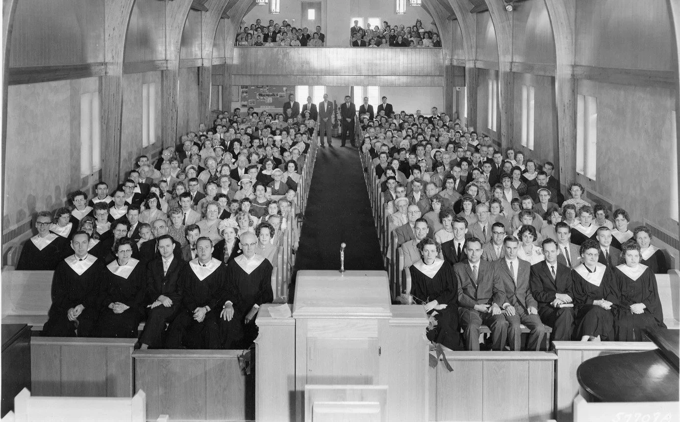 Black and white photo of a large group of people sitting in an auditorium, with some individuals standing at the back and on the balconies. The front row features people in academic gowns and suits, with a podium and microphone in front of them.