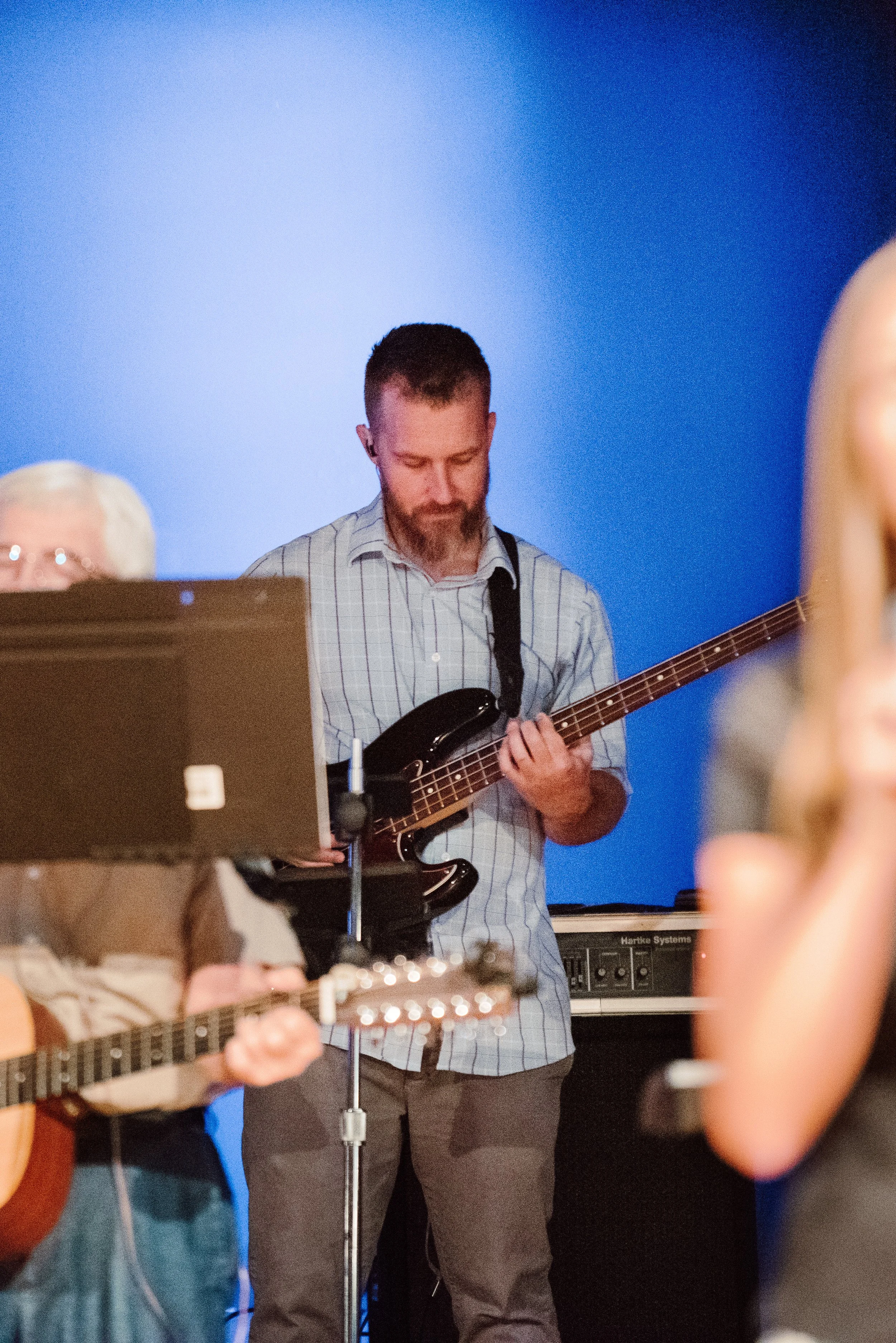 A man with a beard playing an electric bass guitar during a live music performance in front of a blue background, partially obscured by other musicians and instruments.