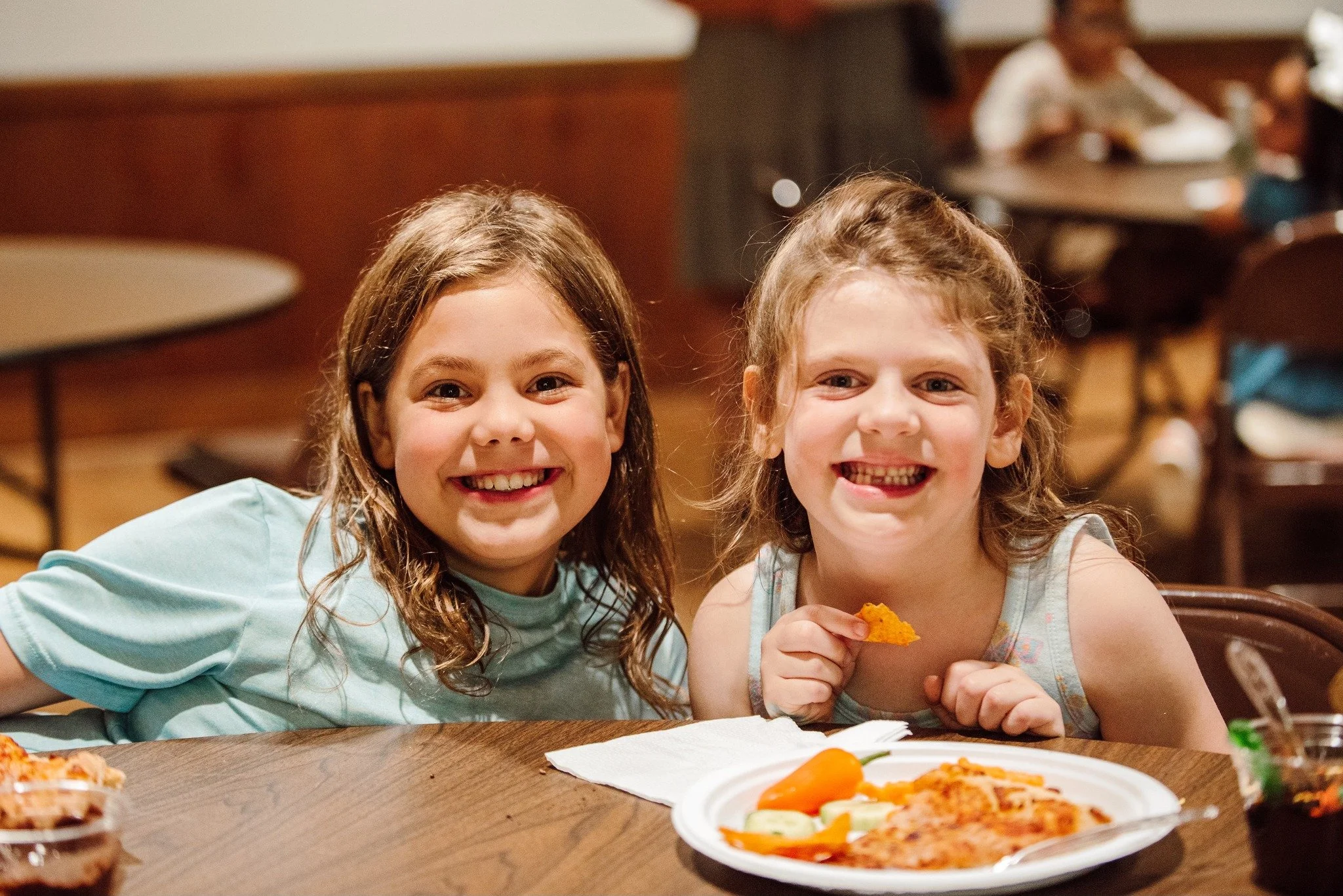 Two smiling young girls sitting at a restaurant table with a plate of pizza, a bowl of sauce, and a napkin, enjoying a meal together.