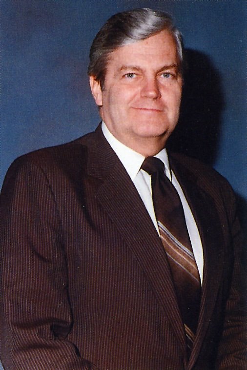 A middle-aged man with gray hair wearing a dark suit, white shirt, and striped tie, standing against a dark blue background.