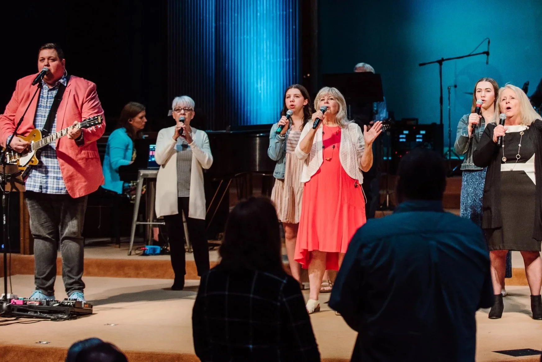 Group of six people performing on stage, singing into microphones, with a pianist playing in the background. The stage has a blue backdrop and a grand piano, with audience members in the foreground.