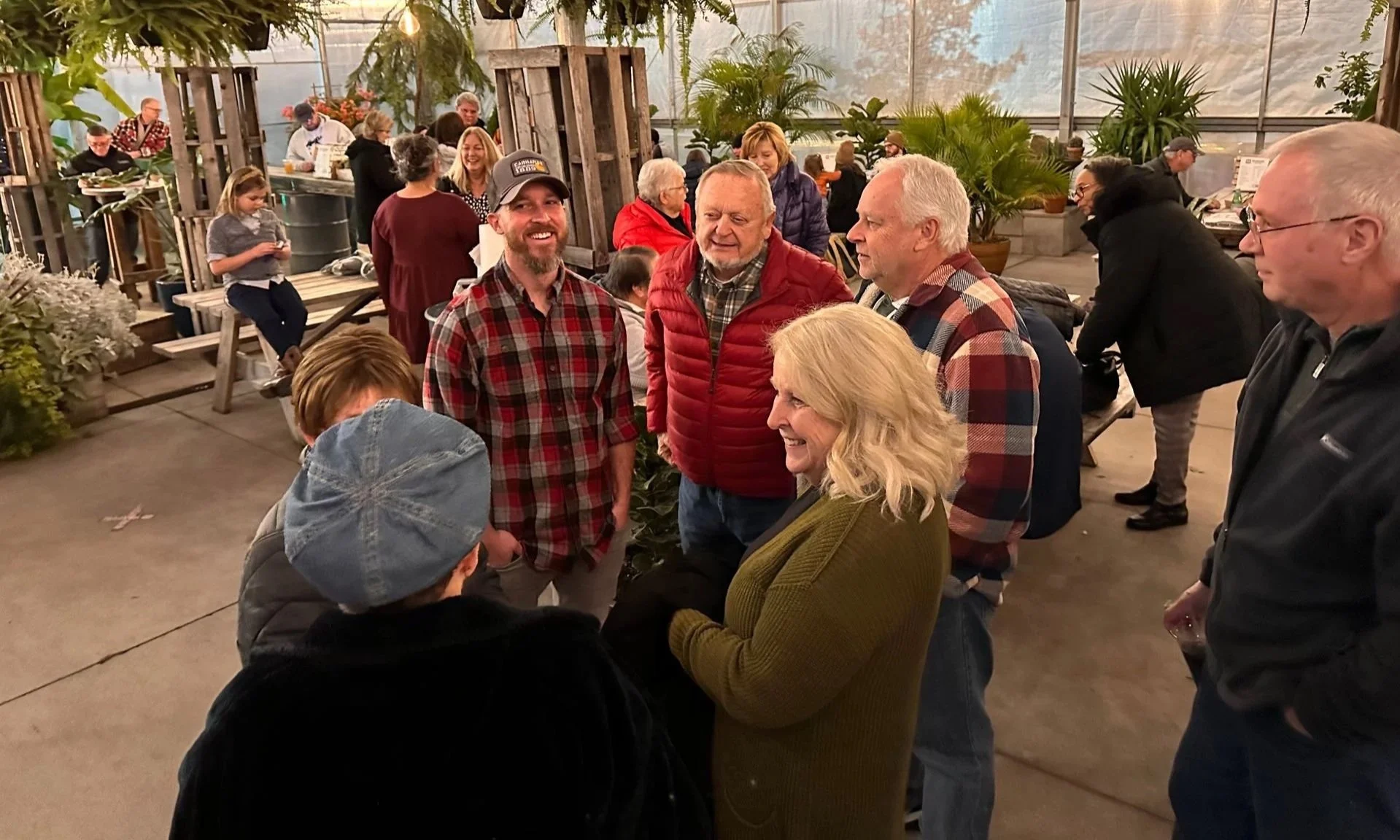 Group of people talking and laughing in a greenhouse-like setting with plants and wooden decor.
