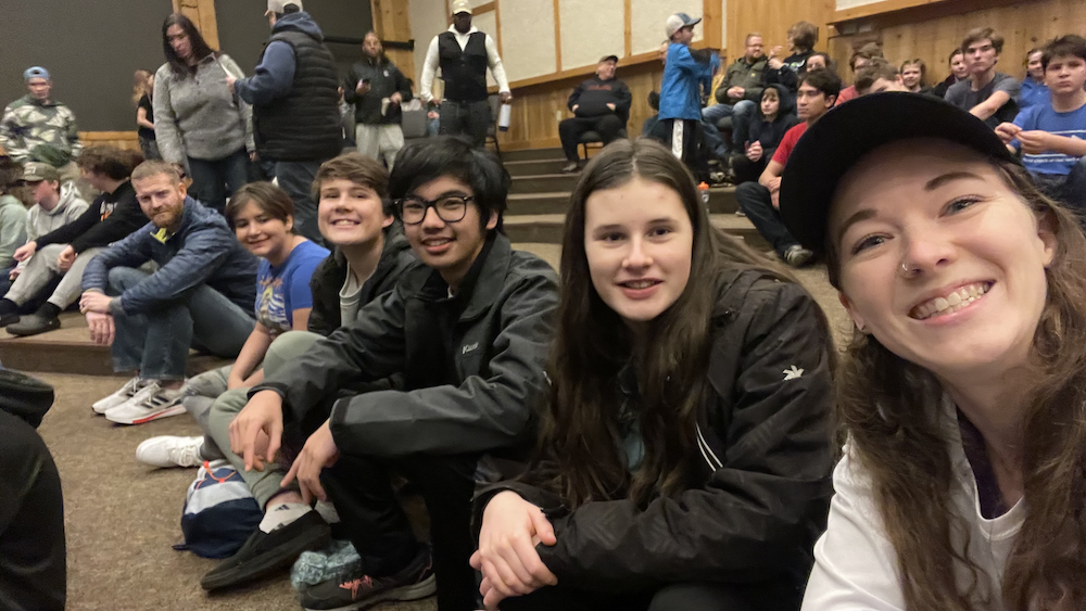 A group of young people sitting on a wooden tiered seating area in what appears to be a theater or auditorium, smiling and looking at the camera. The background shows more people standing and sitting, with some engaged in conversation.