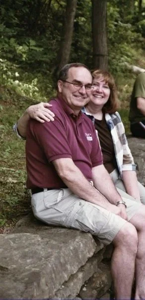 A man and woman sitting on a rock outdoors, hugging and smiling at the camera with trees in the background.