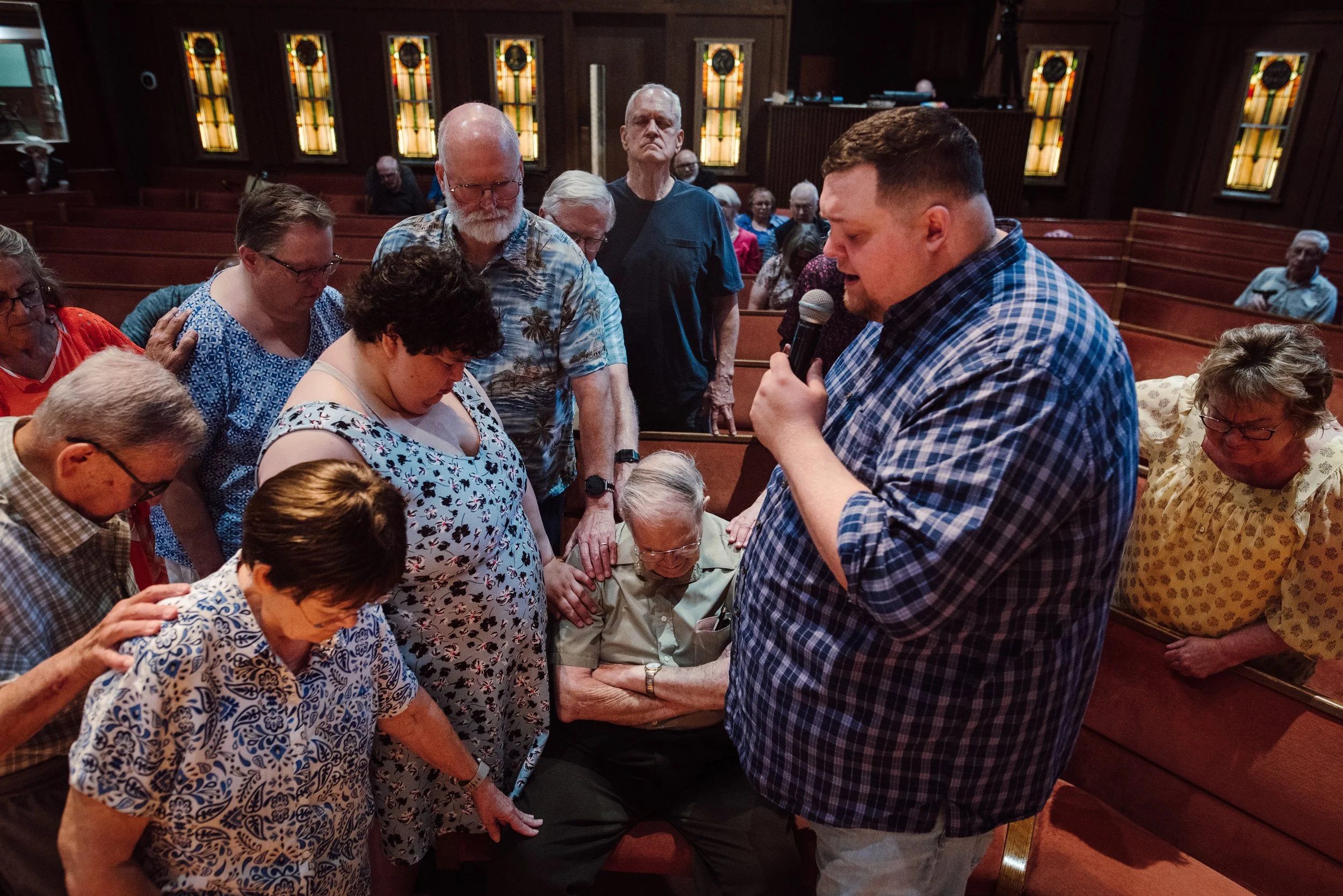 A group of people praying with their heads bowed, including a man holding a microphone, inside a church with stained glass windows in the background.