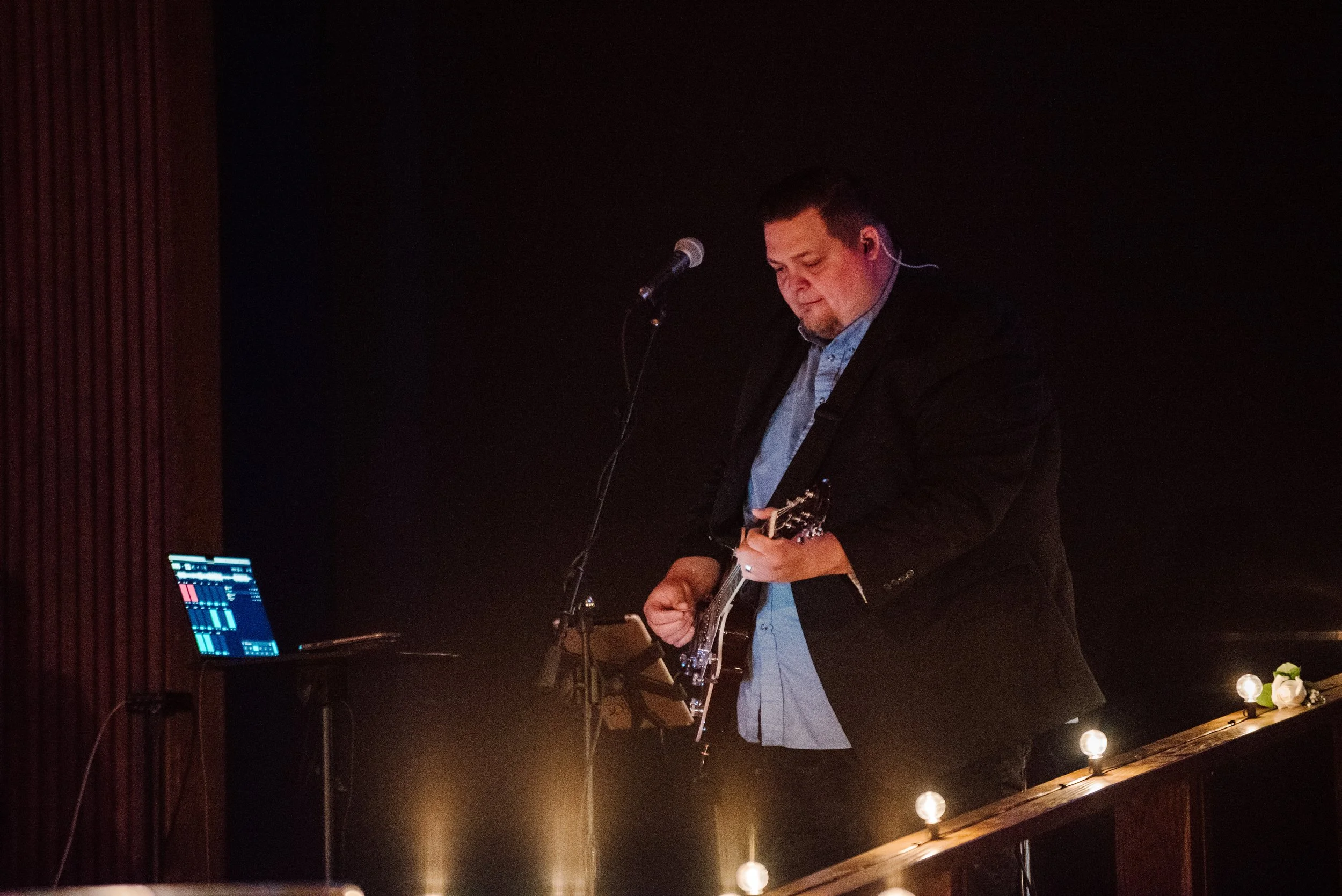 A man in a black suit and blue shirt playing an electric guitar on stage, with a microphone nearby and a laptop on a stand beside him. The stage is dimly lit with warm lighting and string lights at the front.