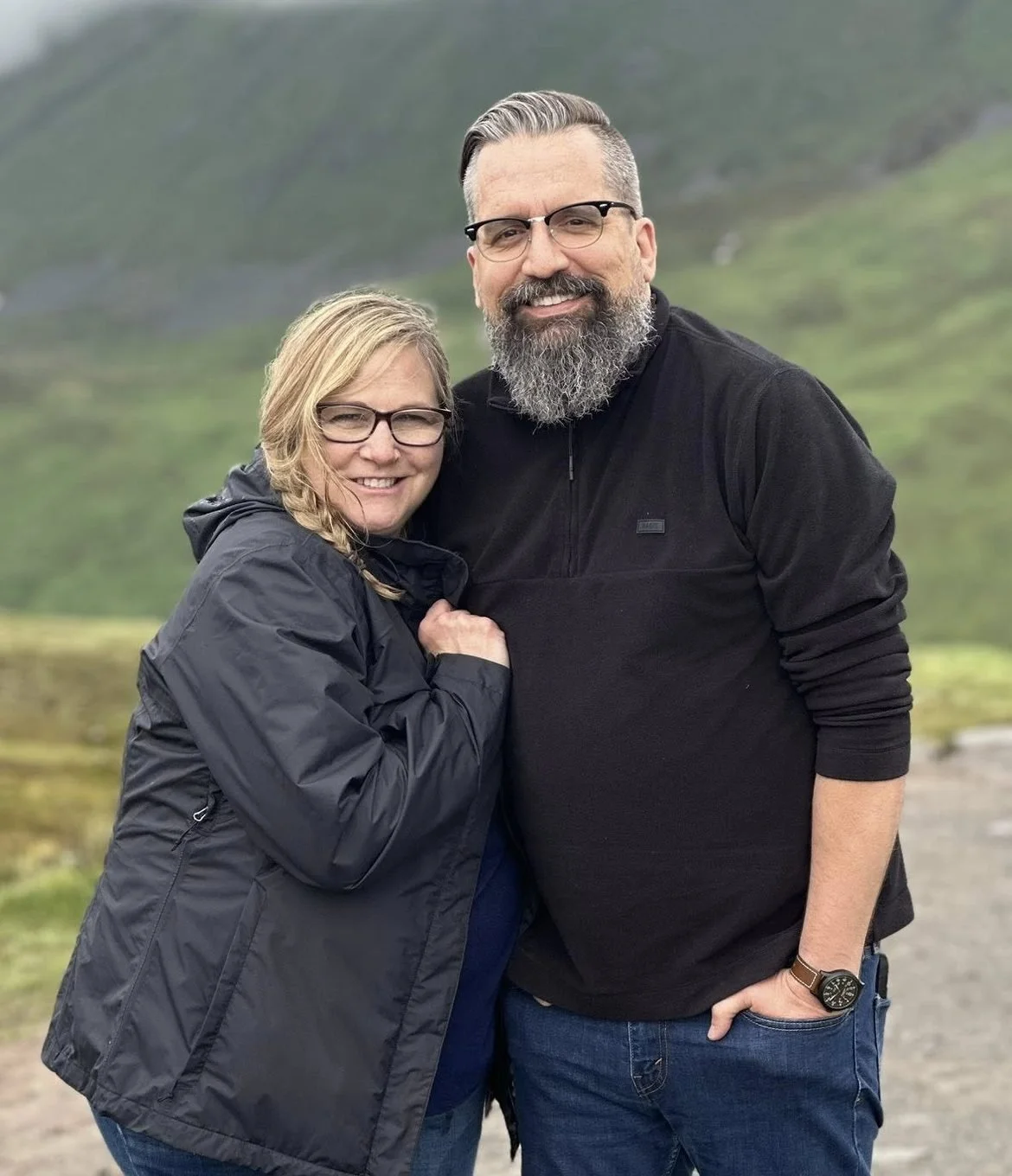 A smiling couple standing outdoors in front of a green mountainous landscape, wearing casual jackets.