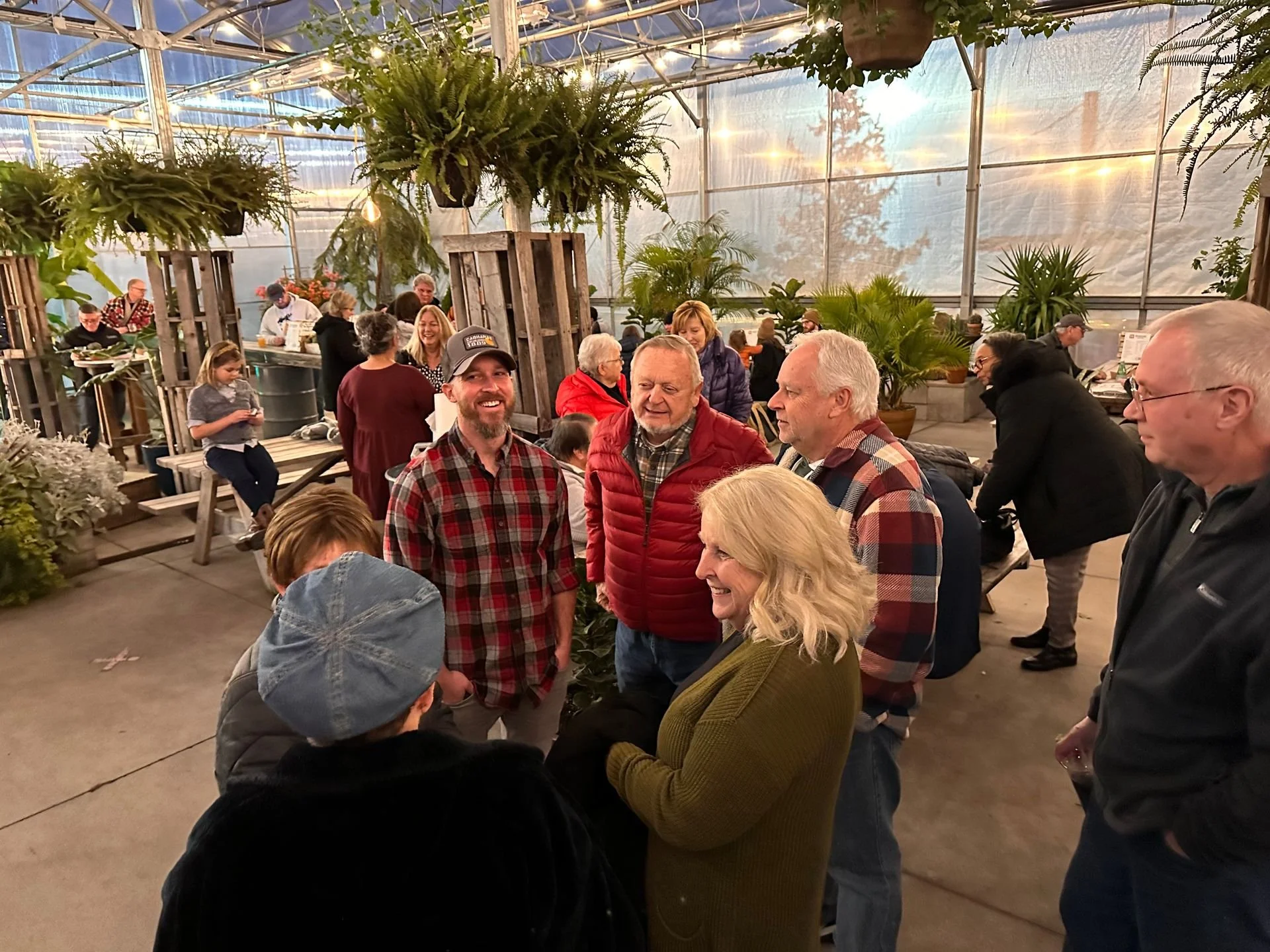 A group of people gathered in a greenhouse-like setting, engaging in conversation and smiling, with plants and other groups of people in the background.