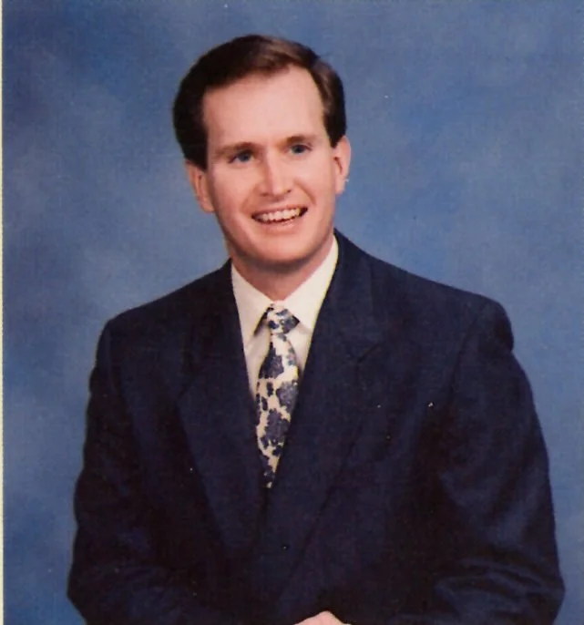 A man with dark brown hair, wearing a navy suit, white shirt, and a patterned tie, smiling in front of a blue background.