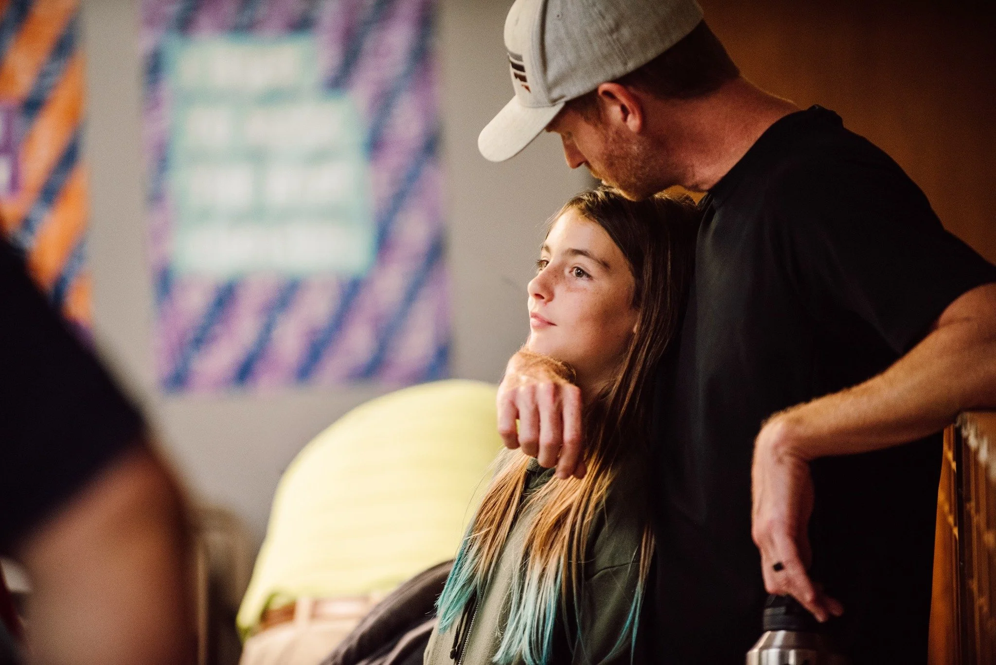 A man with a baseball cap and a girl with long hair, sitting closely together, with the man leaning in and resting his chin on the girl's head, indoors with a colorful abstract background.