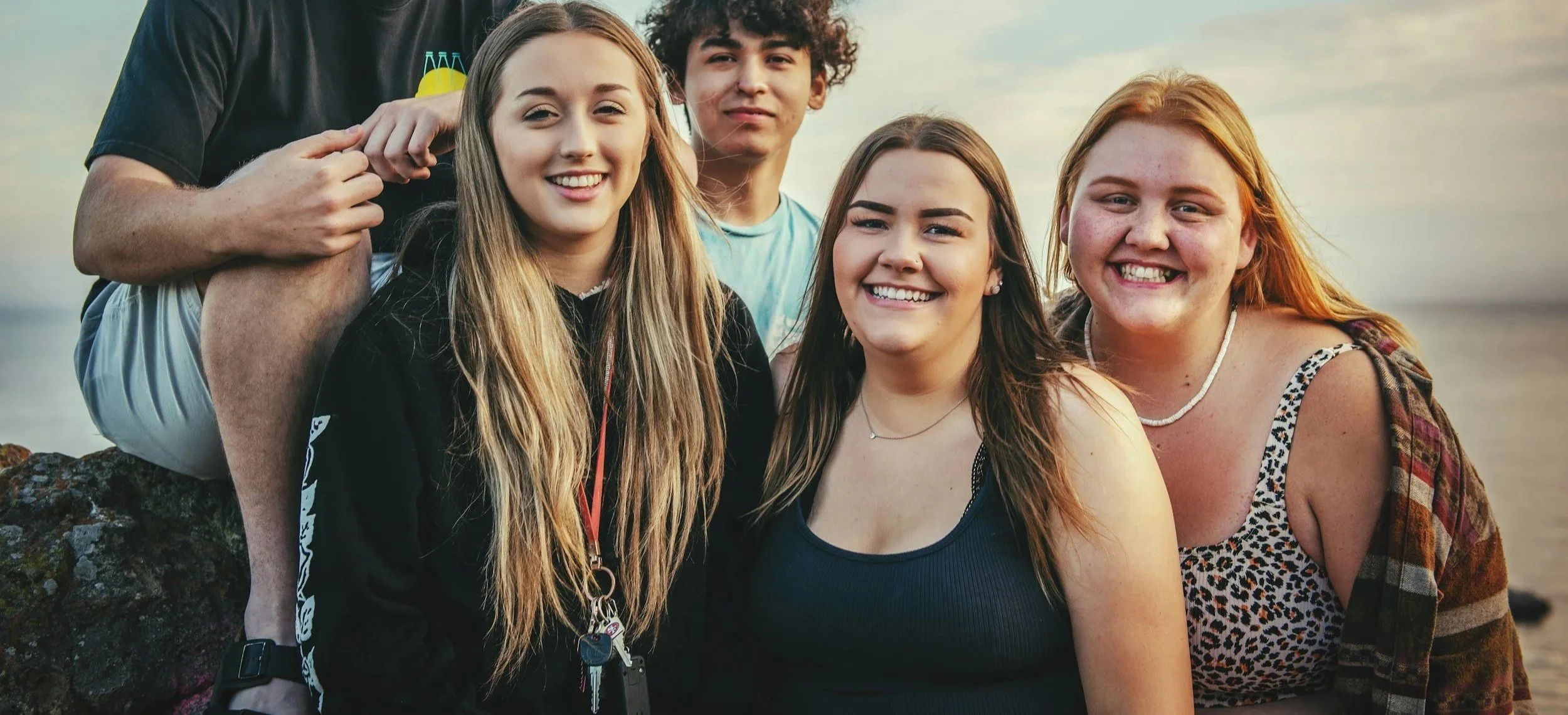 Group of five young friends smiling and posing for a photo by the beach at sunset.