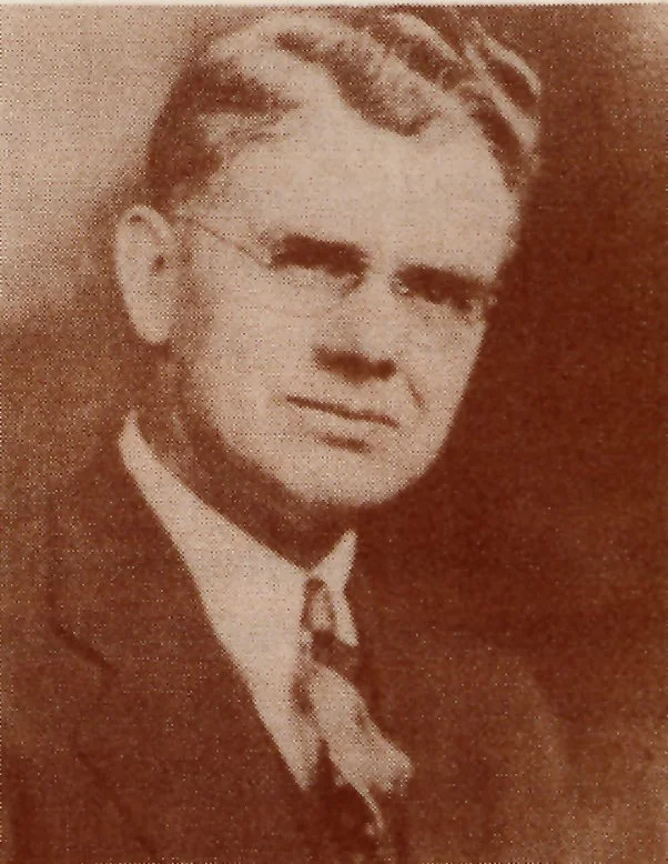 Black and white photo of a man with glasses wearing a suit and tie.