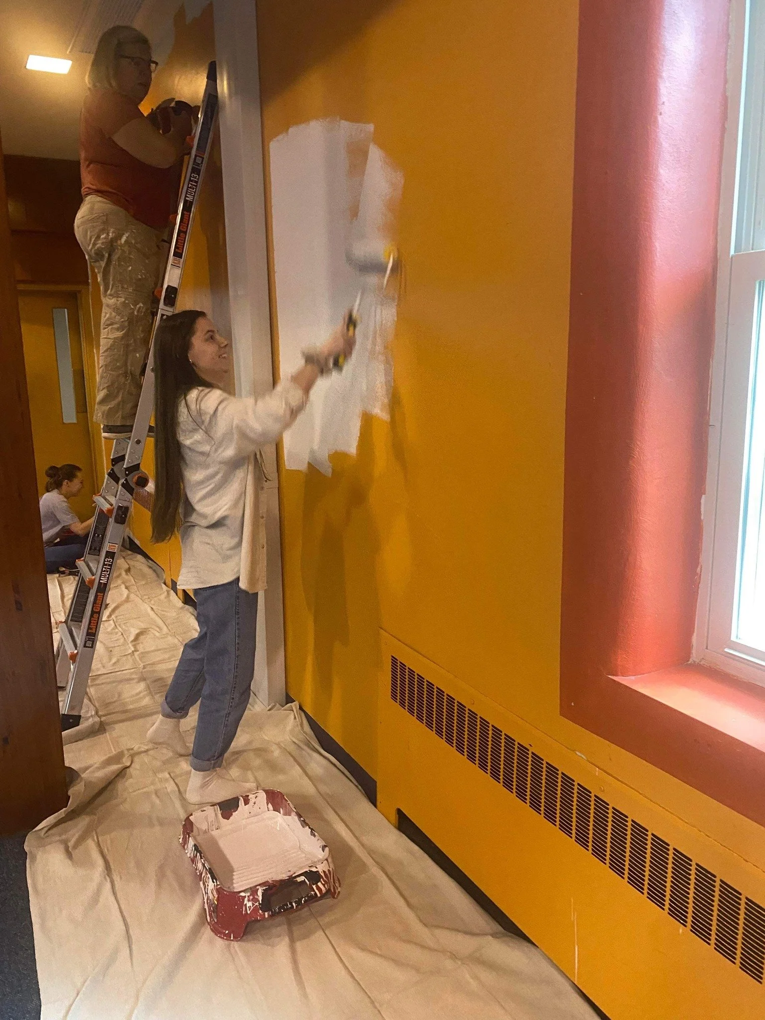 Three women painting a yellow wall in a room. One woman stands on a ladder painting the upper part of the wall, another woman is using a roller to paint the lower part, and a third woman is sitting on the floor, working on a project.