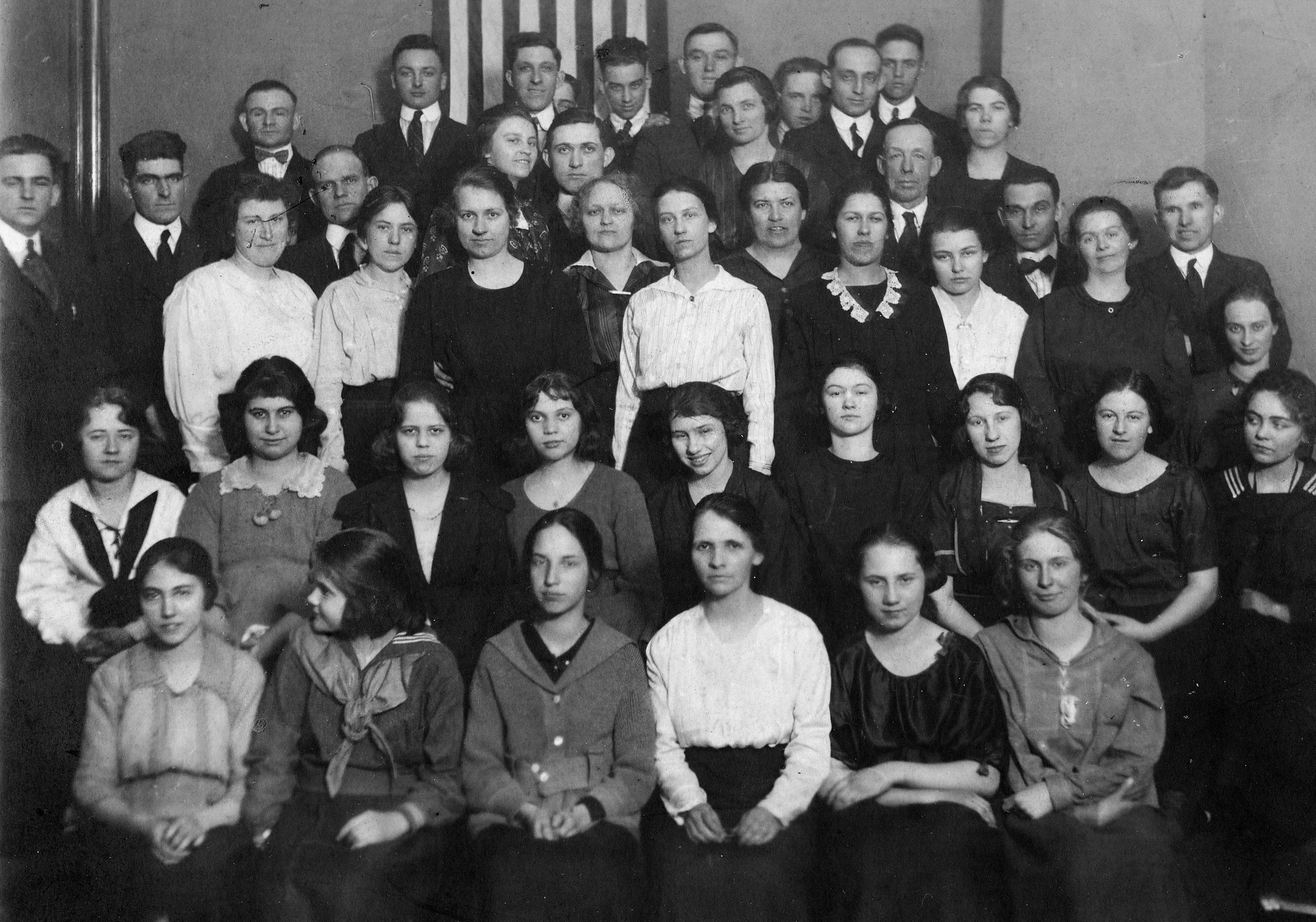A black and white group photo of young men and women, some wearing formal and some casual clothing, gathered indoors against a plain background with vertical stripes.