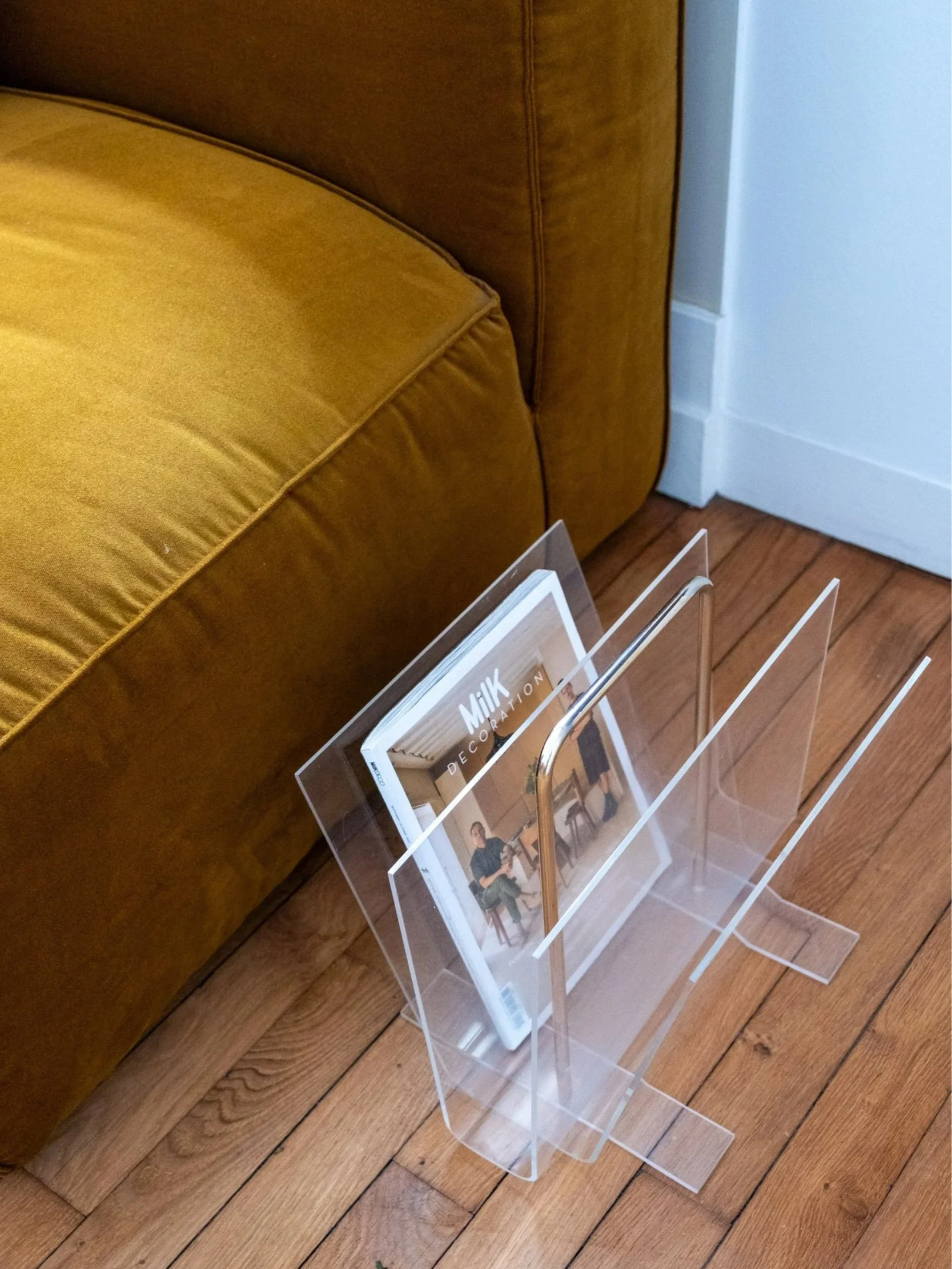 Porte-revues vintage en plexiglas transparent et poignée dorée présenté au pied d'un canapé avec un magazine de décoration