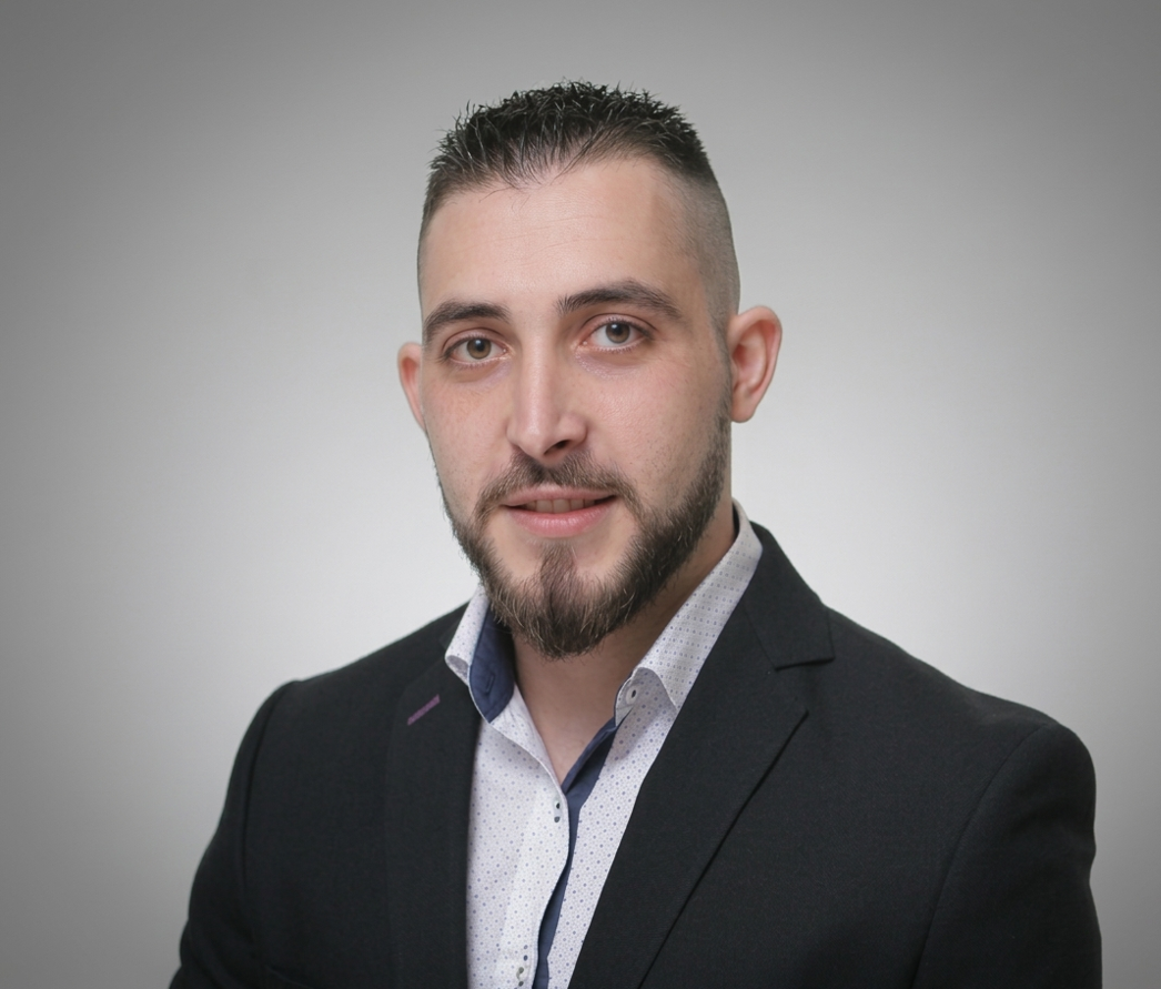 A young man with short dark hair and a beard, wearing a black suit jacket over a white dress shirt, looking at the camera against a plain gray background.