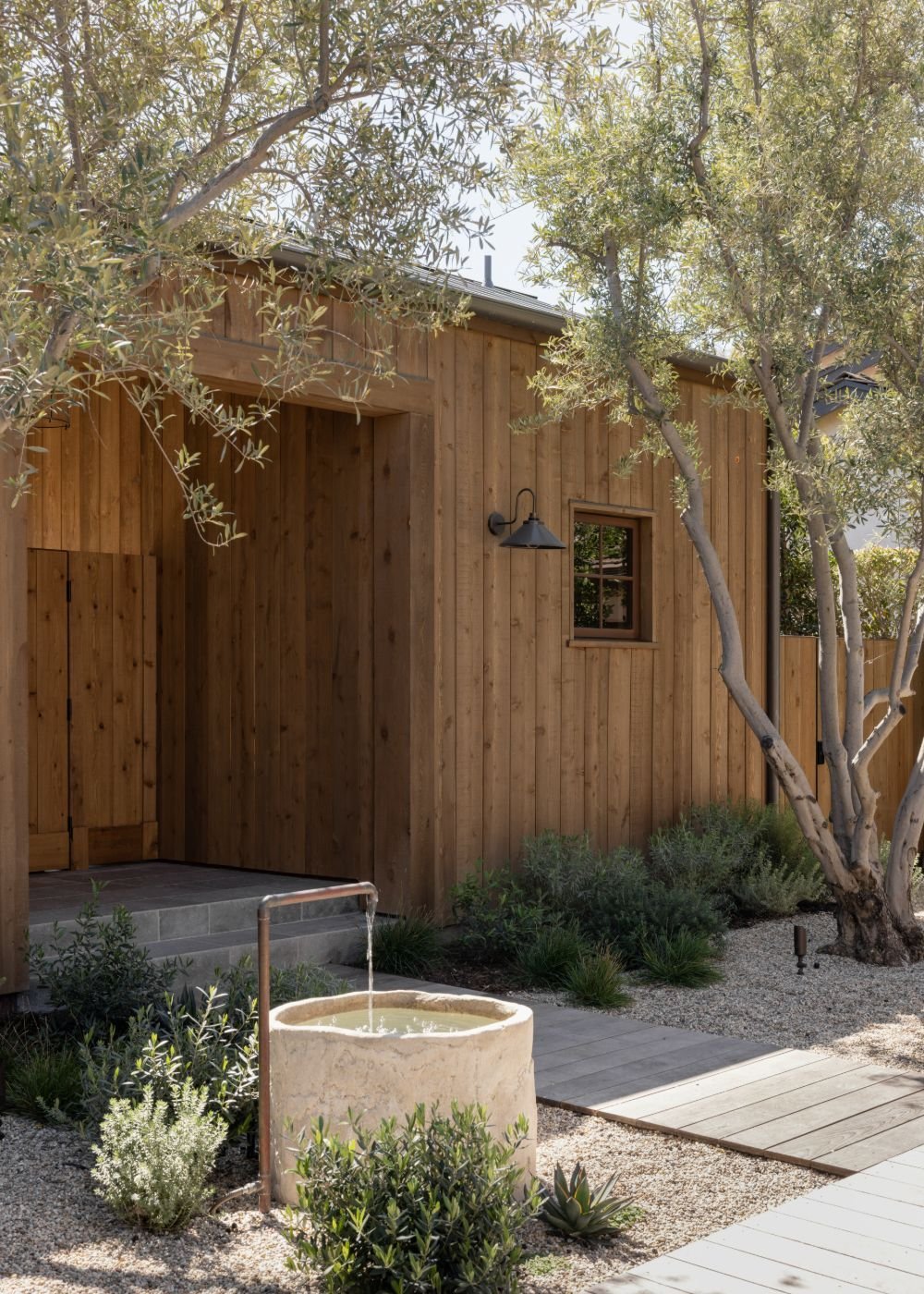 Exterior view of a modern wooden house with small window and outdoor wall lamp, surrounded by trees and desert landscaping including a stone water basin.