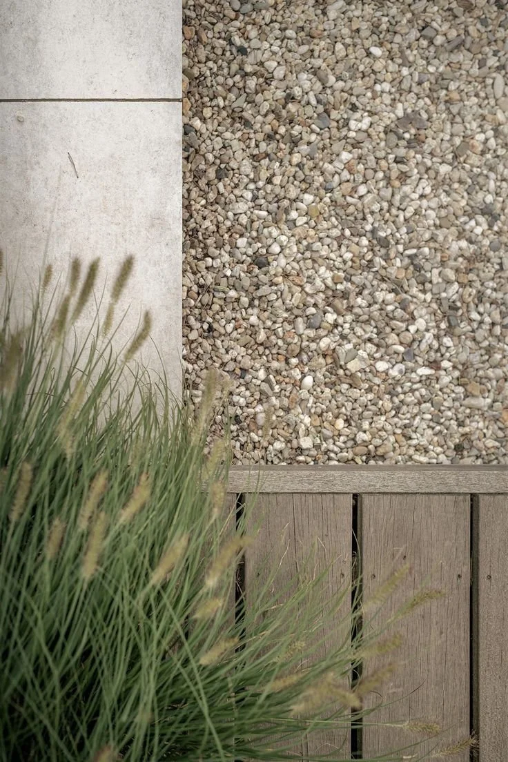 Close-up of a corner of a modern outdoor space with concrete, pebble stones, and wooden flooring, with some grass in the foreground.