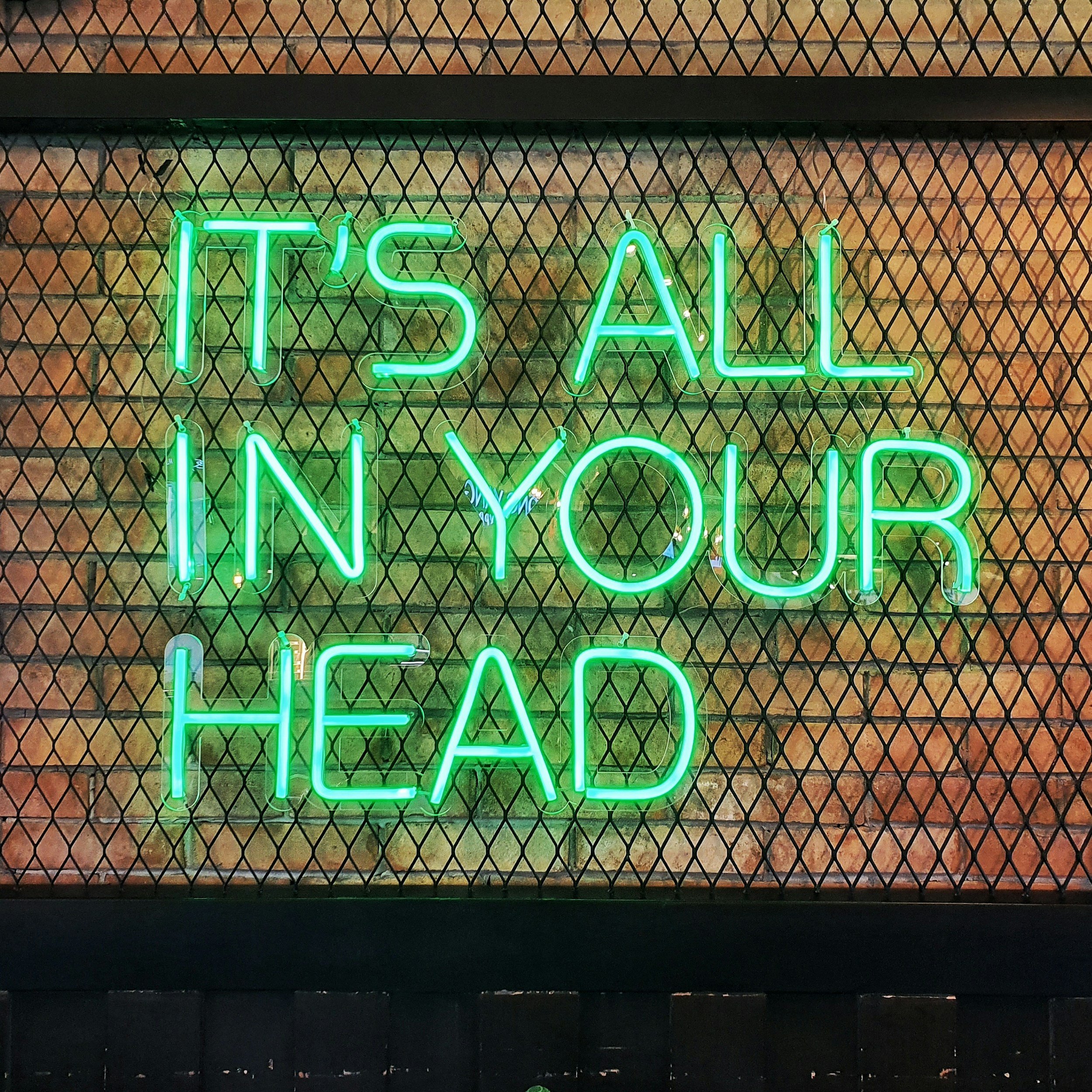 Neon sign that reads 'IT'S ALL IN YOUR HEAD' in green letters against a brick wall background.