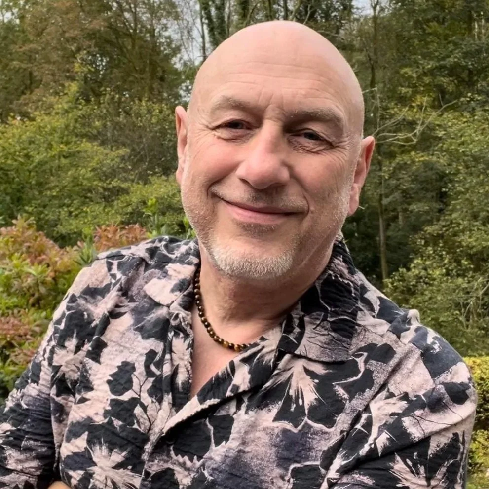 A bald man with a short beard smiling outdoors with trees in the background, wearing a patterned shirt and a beaded necklace.