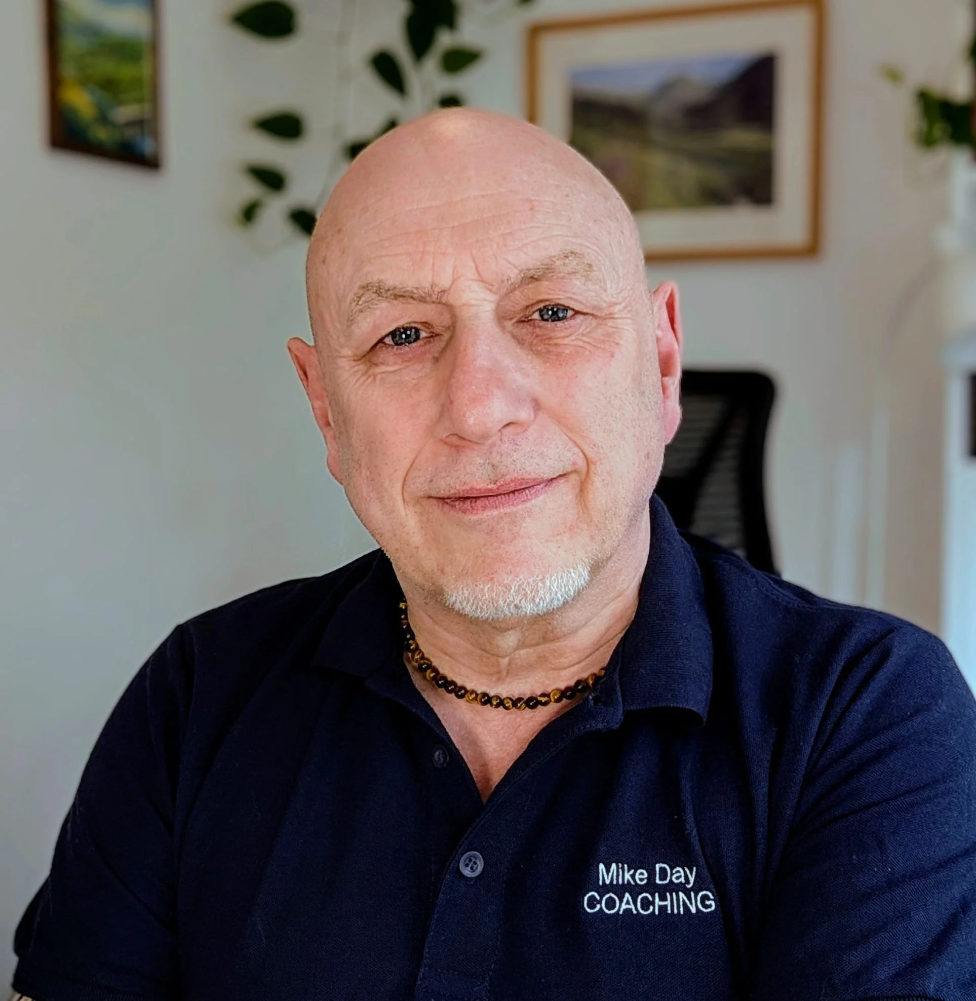 A bald man with a white beard wearing a black shirt with 'Mike Day Coaching' embroidered, sitting indoors in front of a wall with framed pictures and greenery, smiling at the camera.