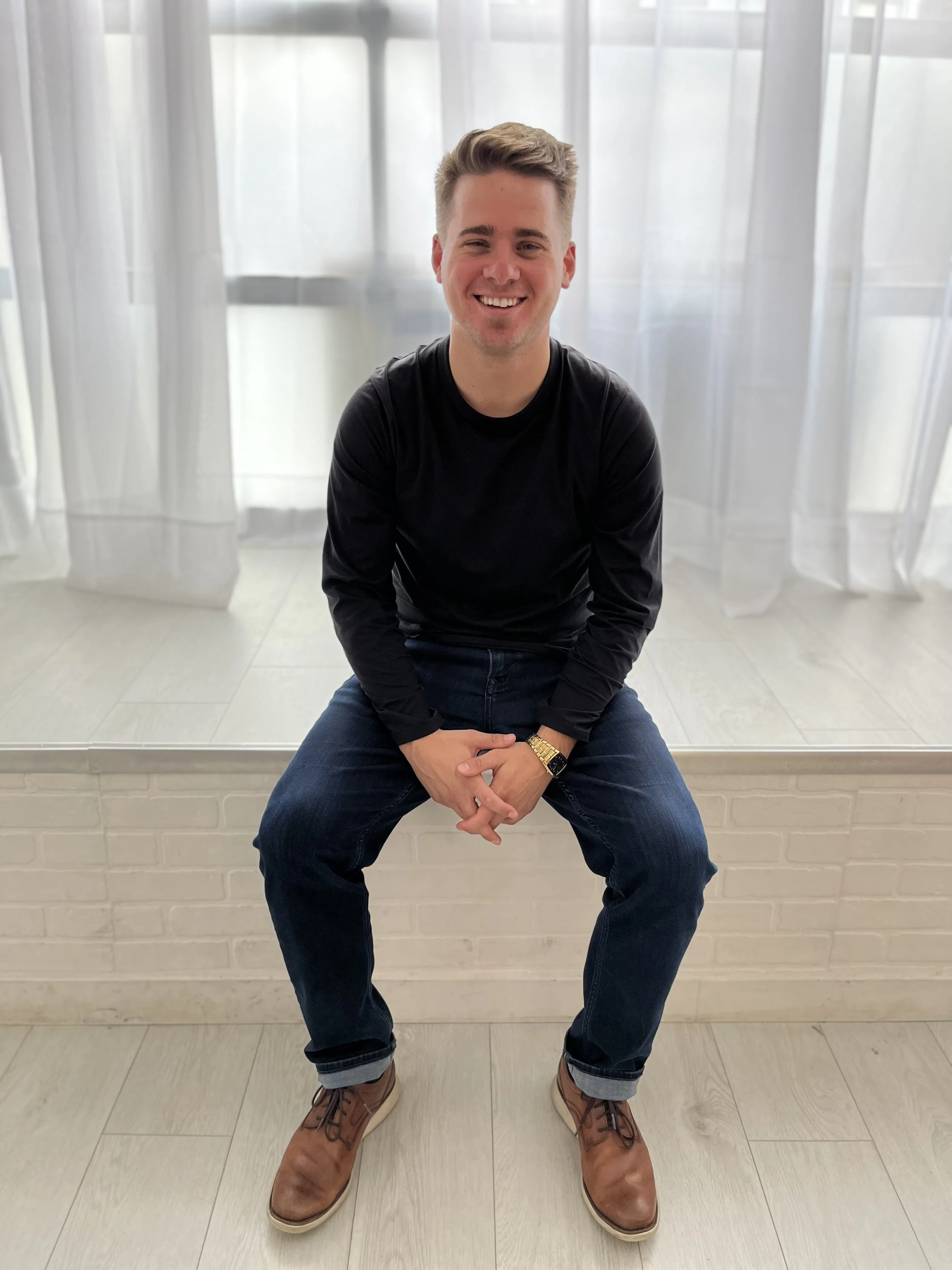 A young man with short, light brown hair, seated on a ledge, smiling at the camera. He is wearing a black long-sleeve shirt, blue jeans with rolled cuffs, brown shoes, a gold watch, and a black wristband. The background features sheer white curtains and a light-colored tiled floor.