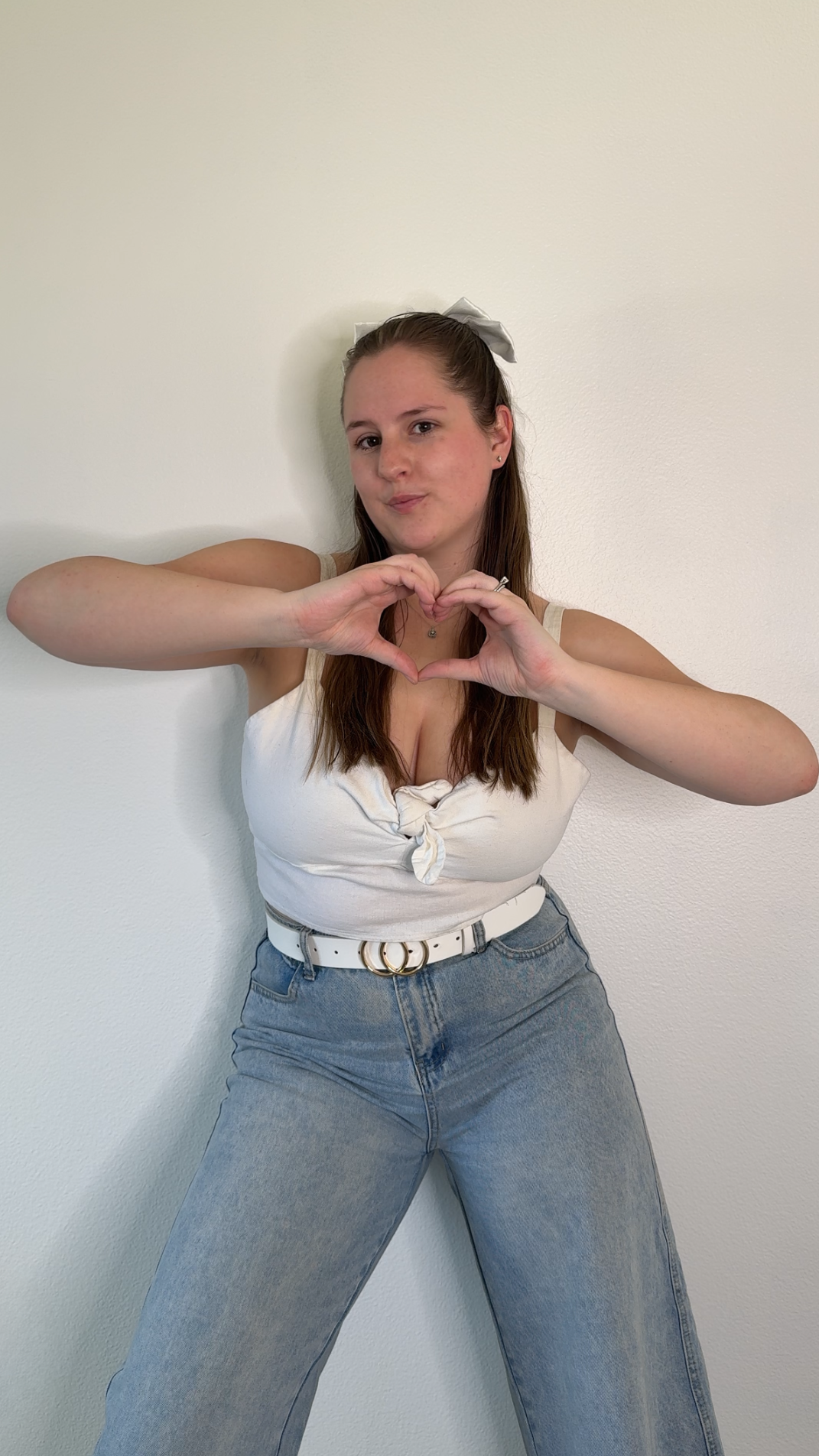 A young woman with long brown hair, wearing a white tank top with a front knot and light-wash jeans, standing against a plain white wall. She is making a heart shape with her hands and looking at the camera.