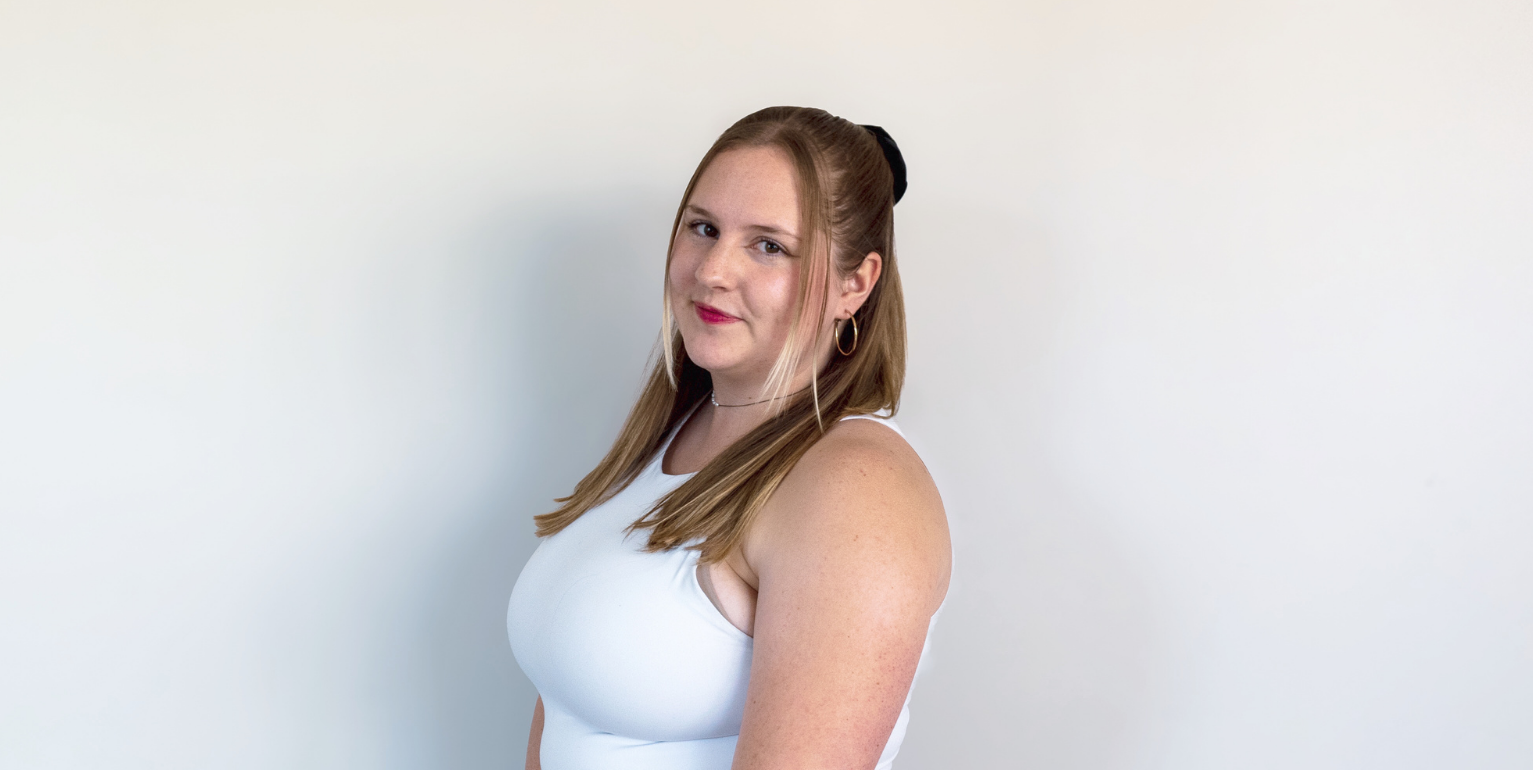 A young woman with long light brown hair, wearing a white sleeveless top, standing against a plain white wall.