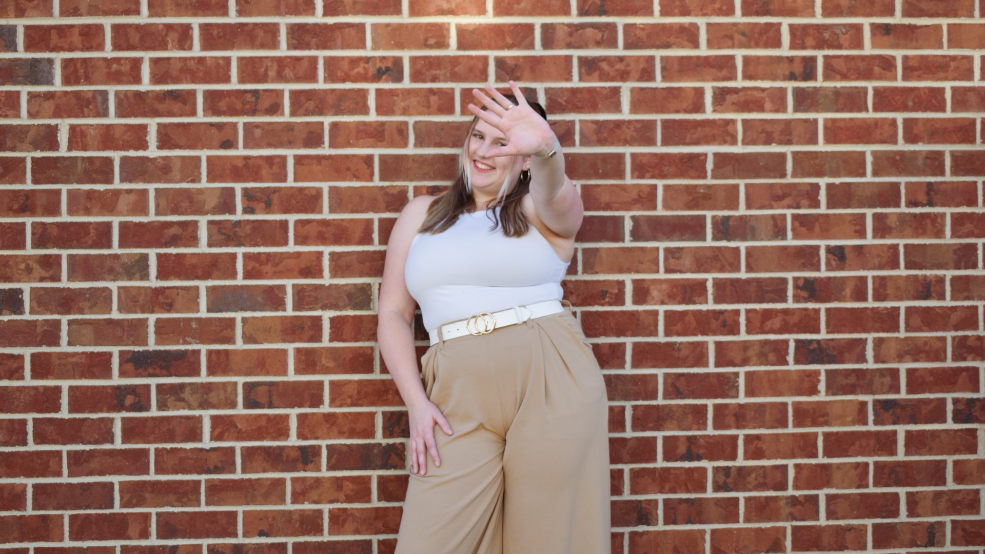 A woman standing against a brick wall, smiling, with her left hand raised and her right hand resting on her hip.