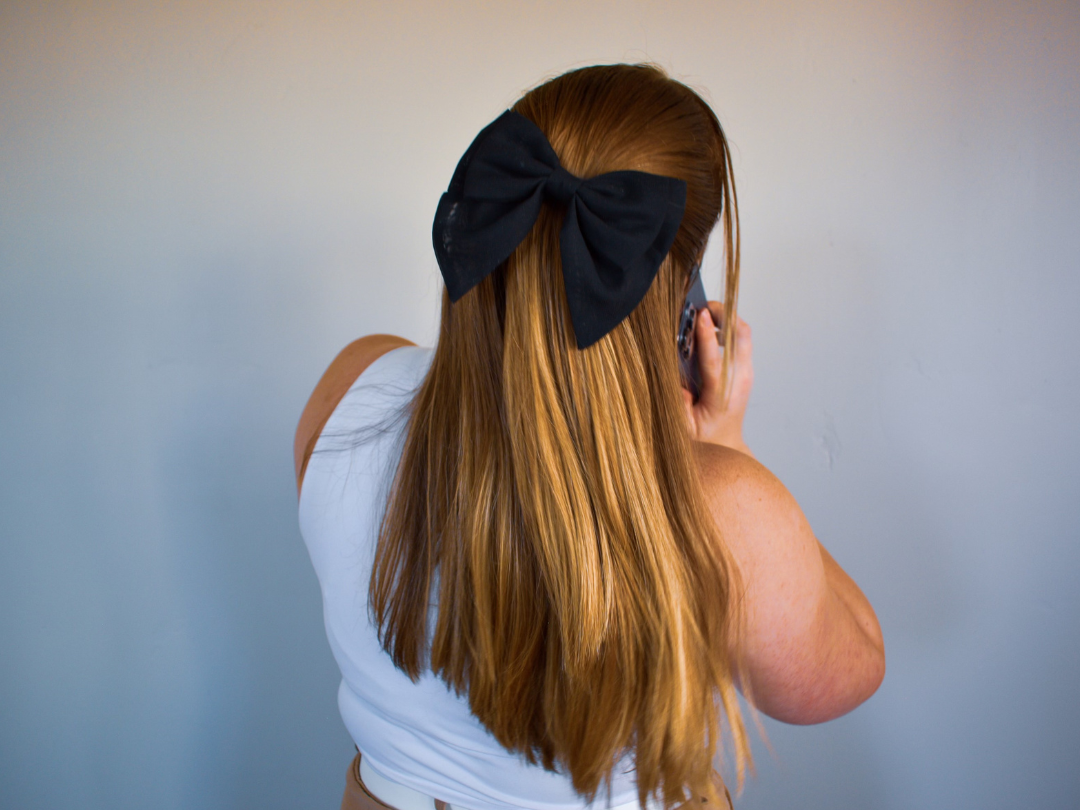 A woman with long, straight red hair, wearing a large black bow on her head, is holding a phone to her ear against a plain gray wall.