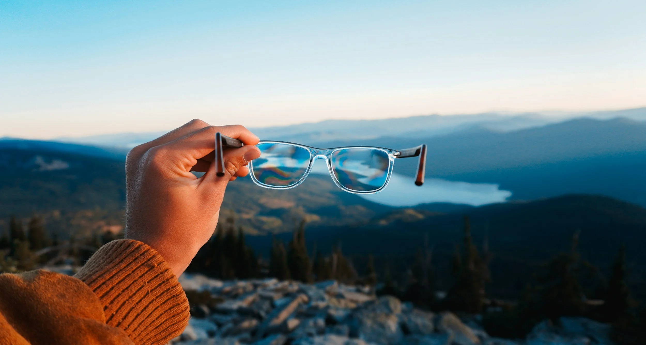 A person holding a pair of glasses in front of a mountain landscape with blue sky and distant hills.