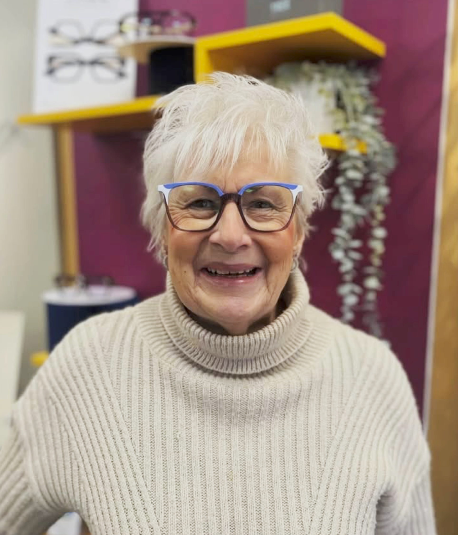 Close-up of a happy customer, wearing her new glasses at the practice. Display walls featuring new frames feature in the background.