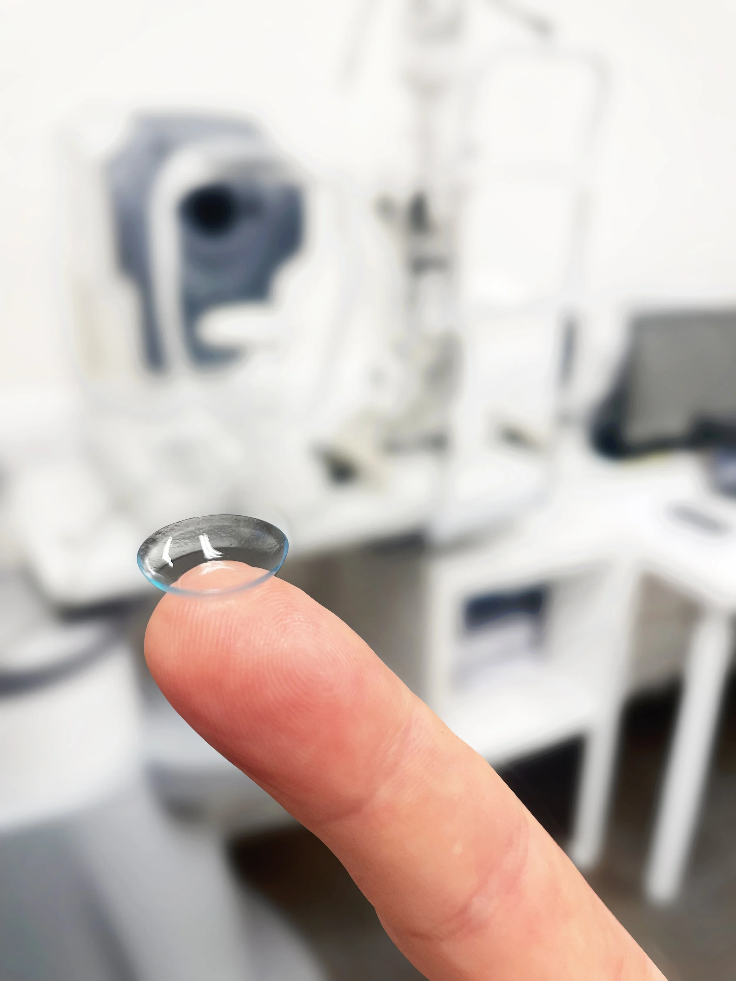 Close-up of a finger holding a small, transparent contact lens in front of a blurry background of machines.