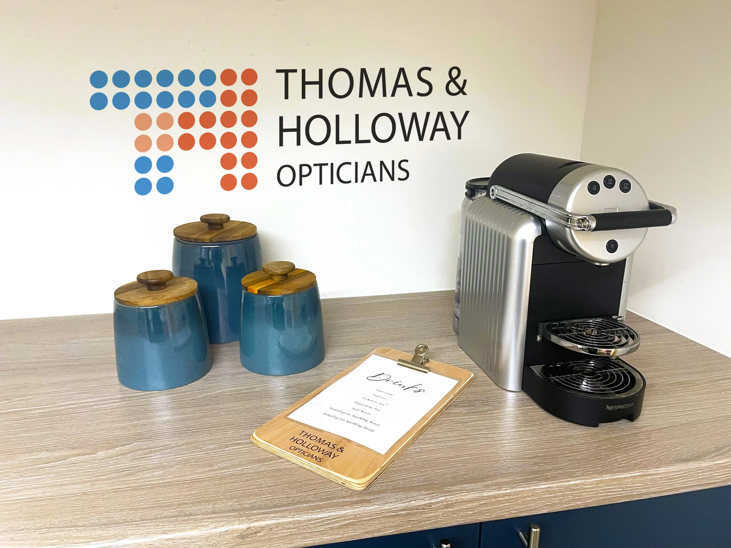 An office reception area with a sign reading 'Thomas & Holloway Opticians', a blue cup with wooden lids, a coffee machine, a clipboard with a menu, and a small blue stool.