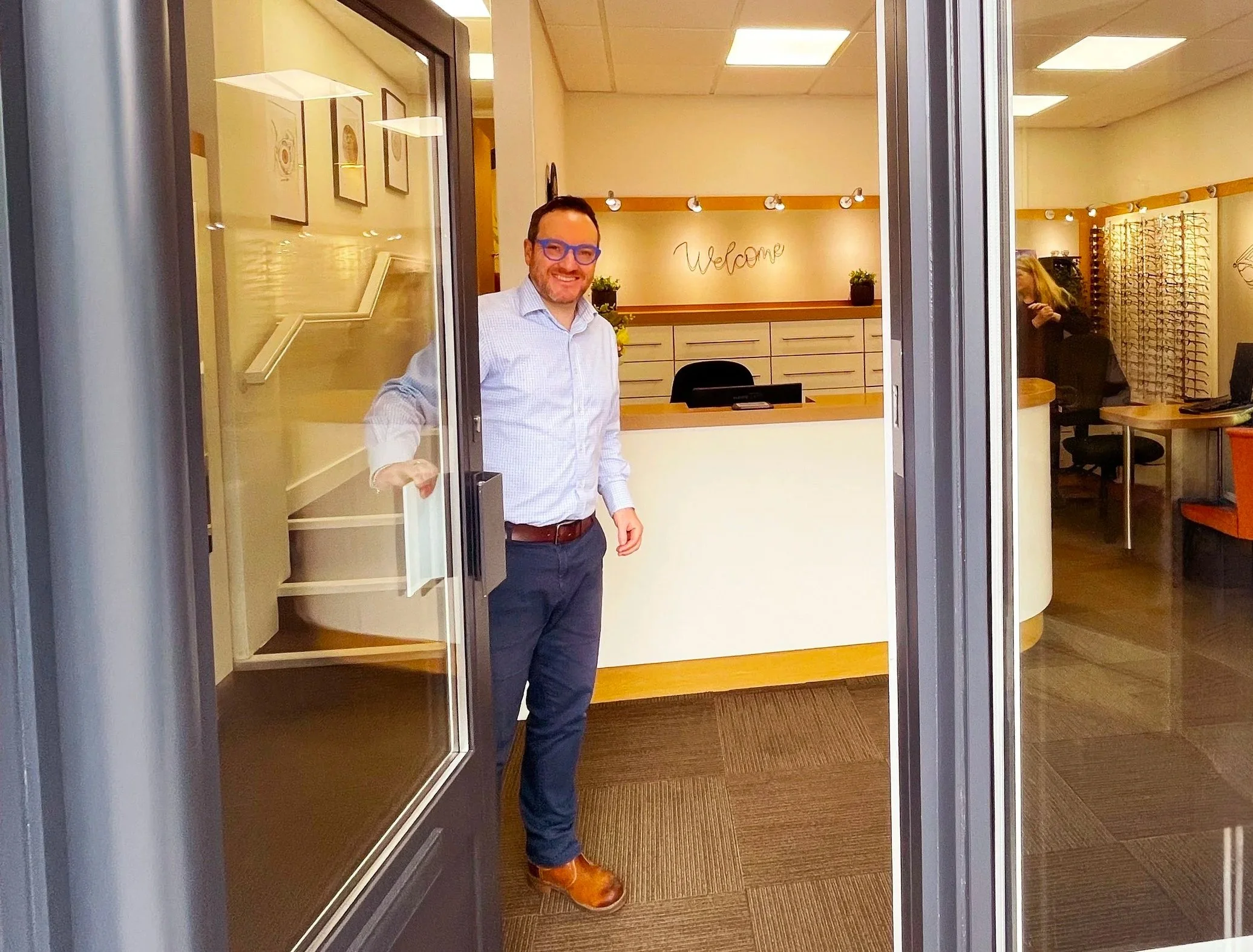 Mark Holloway stands at the main practice entrance, smiling, inside a well-lit reception area with a welcome sign on the wall.