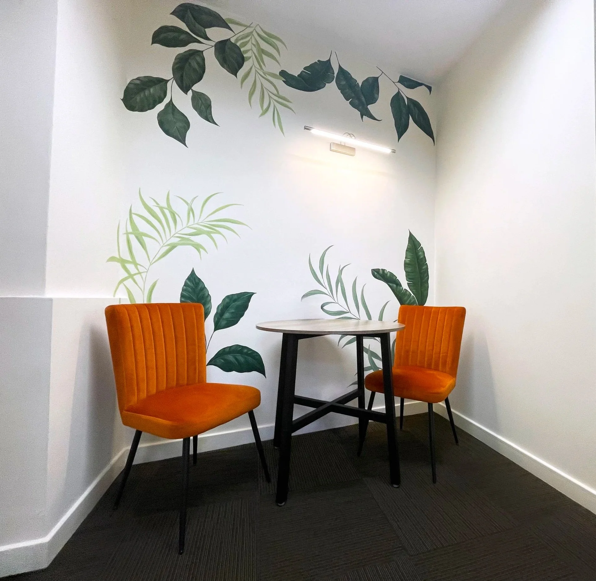 A consultation area within the practice. A corner of a room with a round table and two orange velvet chairs. The wall behind has a leaf-patterned mural with green and dark green leaves.