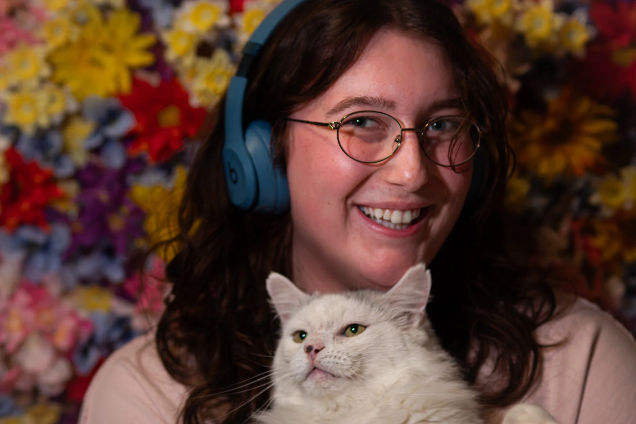 A smiling woman with glasses wearing blue headphones holds a white cat with green eyes in front of a colorful flower background.