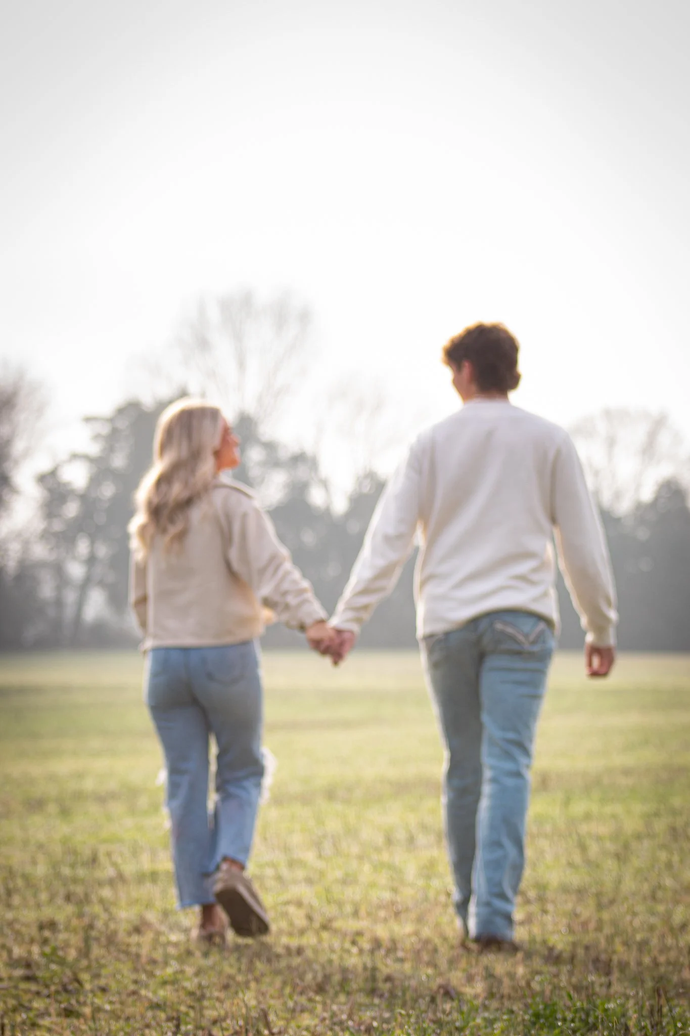 A couple holding hands and walking in an open grassy field, with trees in the background during daytime.
