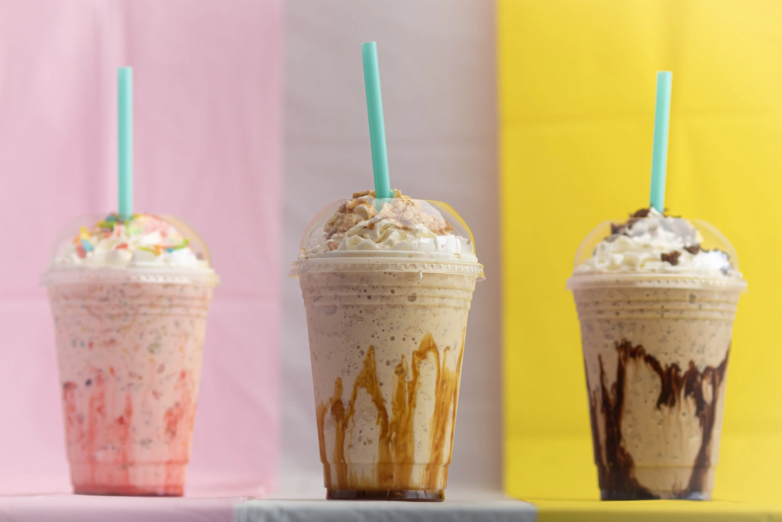 Three milkshakes in clear plastic cups with dome lids and blue straws, each topped with whipped cream and sprinkles or cookie crumbs, set against pink, white, and yellow backgrounds.