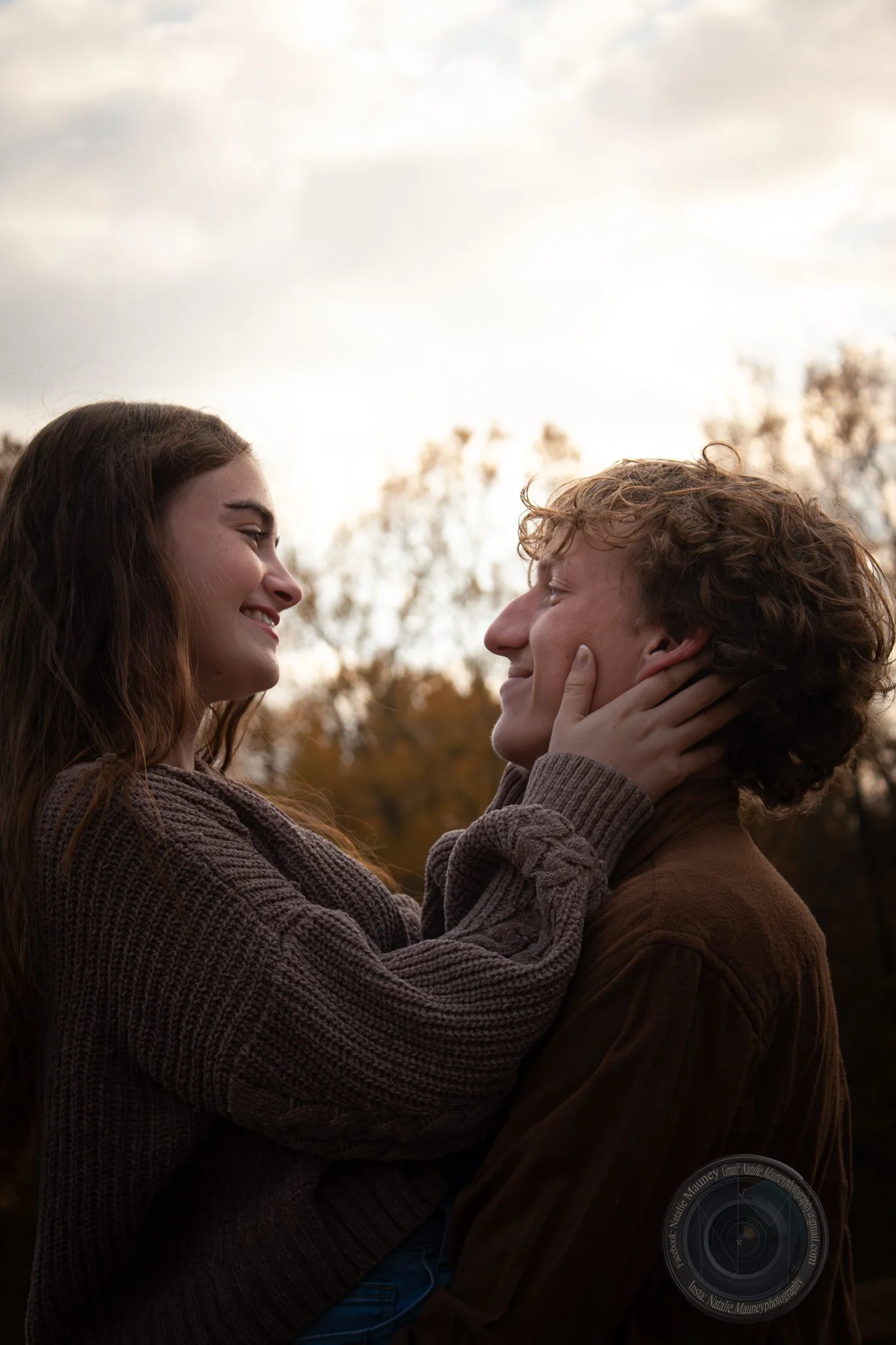 A couple sharing a tender moment outdoors, with the woman holding the man's face and smiling at him as the sun sets behind trees.